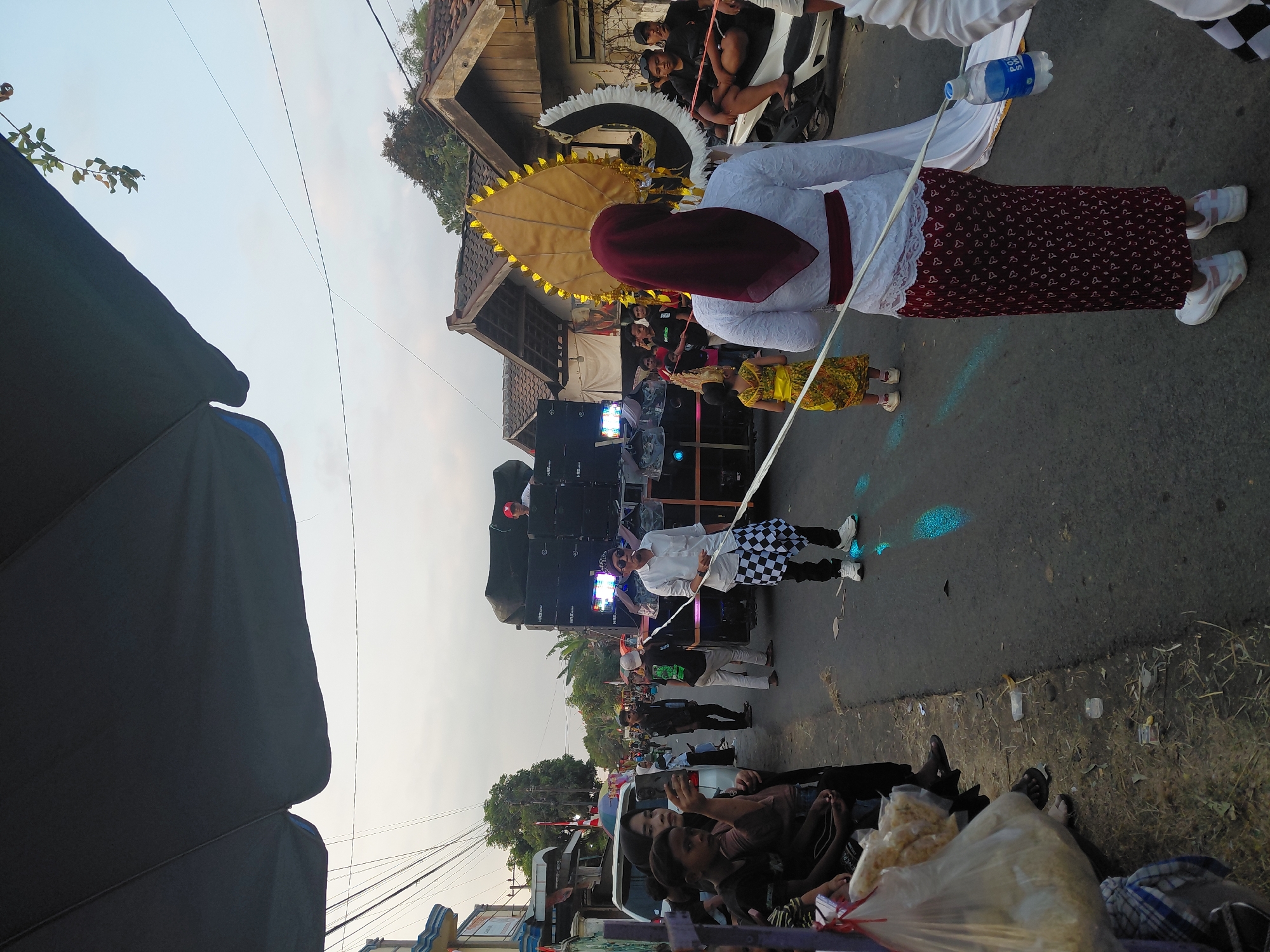 In Bimo, Indonesia, on an outdoor street, a festive gathering is taking place. A large sound system is set up on the back of a truck, with speakers and a screen visible. Several people are gathered around, some watching the events unfold, while others are involved in the proceedings.
In the foreground, a woman wearing a white traditional top, a maroon headscarf, and a patterned red skirt stands with her back to the camera, holding a white fabric. To her left, a young girl in a yellow and gold traditional dress also stands facing away. Further left, a group of people, including men and children, are seated or standing by the side of the road, some holding phones and looking towards the truck. A large plastic bag filled with snacks is placed on the ground near them.
A man in a white shirt and checkered pants, wearing sunglasses and a red cap, stands in the middle of the street, holding a white ribbon or streamer. He appears to be participating in a procession or performance. Other individuals are scattered throughout the scene, some appearing to be part of the sound system crew or performers. The environment suggests a daytime event, with the sky overcast and houses lining the street in the background. The overall impression is one of a community celebration.