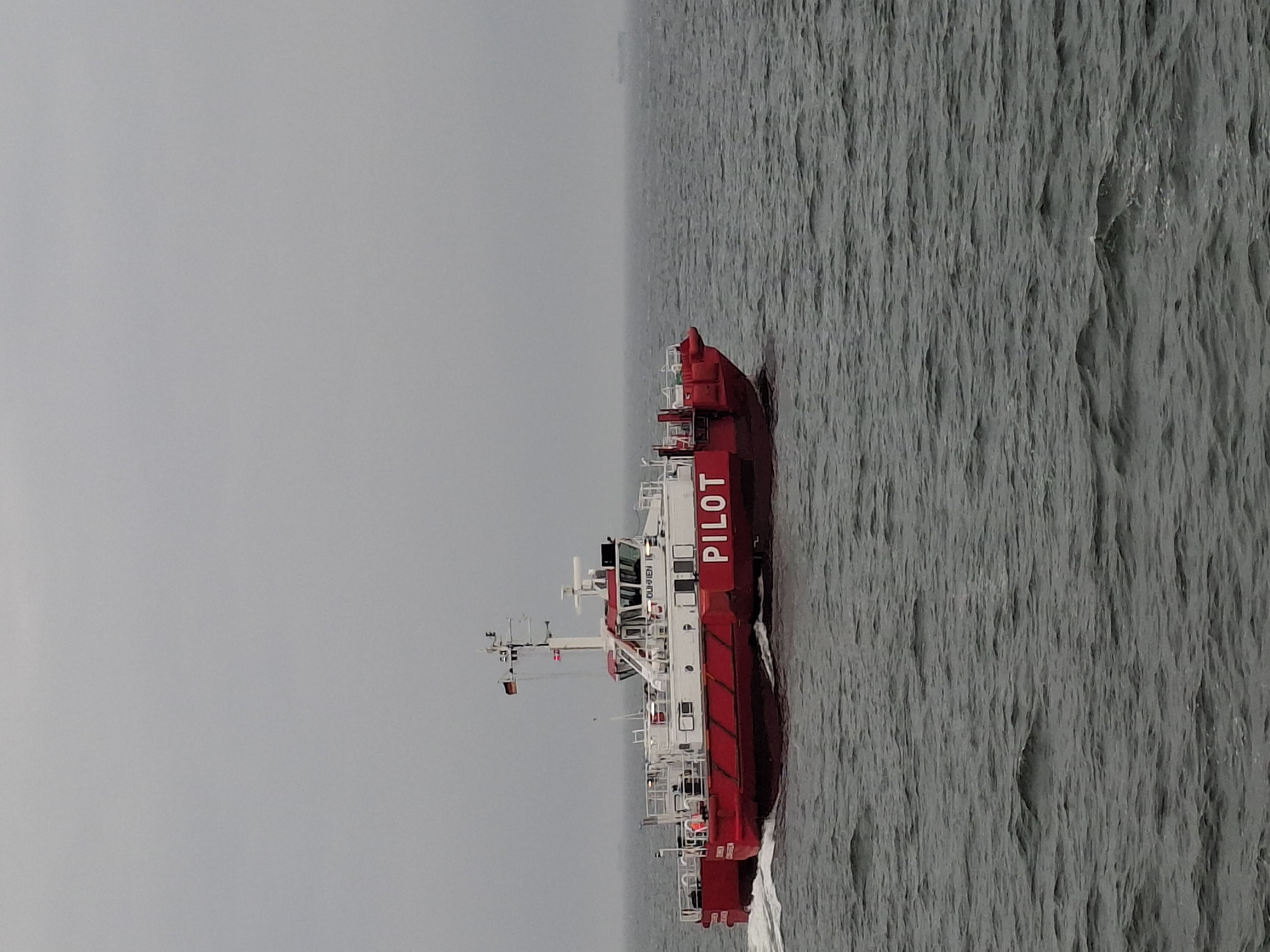 A red and white pilot boat with the word "PILOT" in large white letters on its side is traveling through the water. The water is gray and choppy. The sky is overcast and gray. The pilot boat is in the city of Cuxhaven, Germany.