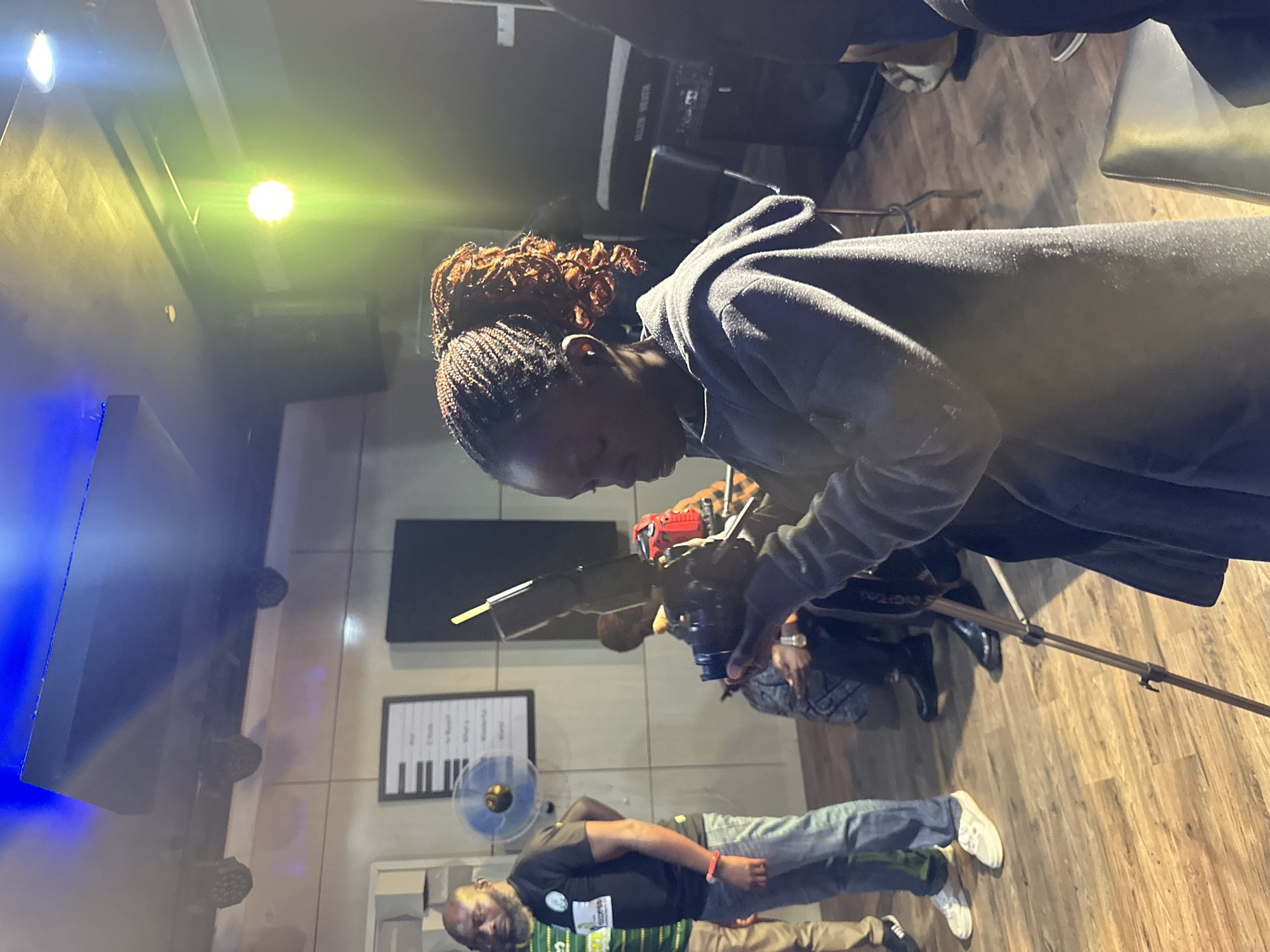 The image shows a group of people in what appears to be a studio or performance space with wooden flooring and a textured wall. The lighting is dramatic, with bright blue and yellow spotlights illuminating the ceiling. In the foreground, a young woman with braided hair is focused on operating a video camera mounted on a tripod. She is wearing a gray hooded jacket. To her left, a man in a black t-shirt and jeans is standing, and further to the left, another person in a green and white striped shirt is partially visible. In the background, there are musical instruments, including what looks like a keyboard on a stand, and a framed item resembling a musical score on the wall. There is also a blue fan visible on the wall. The overall atmosphere suggests a creative environment, possibly for recording music or filming content. The specific location in Barakin Lerawi, Nigeria, is not explicitly indicated by any visible cues in the image.
