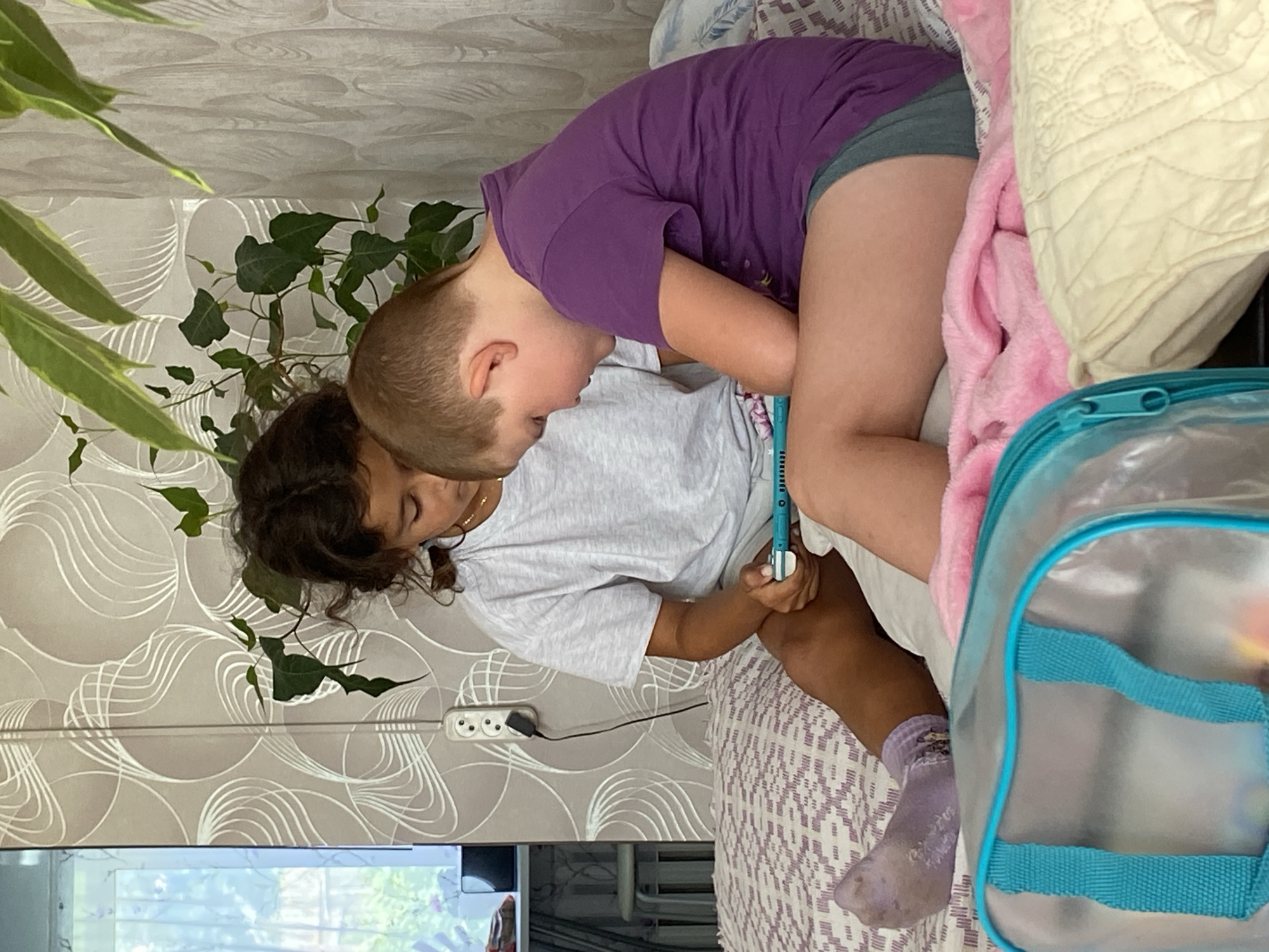 In a domestic indoor setting, two children are seated on a bed, engrossed in a portable electronic device. The child on the left, with curly dark hair, wears a light grey t-shirt and shorts, with a purple sock on their left foot. The child on the right, with short blonde hair, is dressed in a purple t-shirt and grey shorts. A pink blanket and a clear plastic bag with turquoise trim are visible in the foreground. The background features patterned wallpaper and a plant with green leaves. A wall socket with a plug is also present. The image appears to be taken in a bedroom. The location is identified as Otchor, Russia.