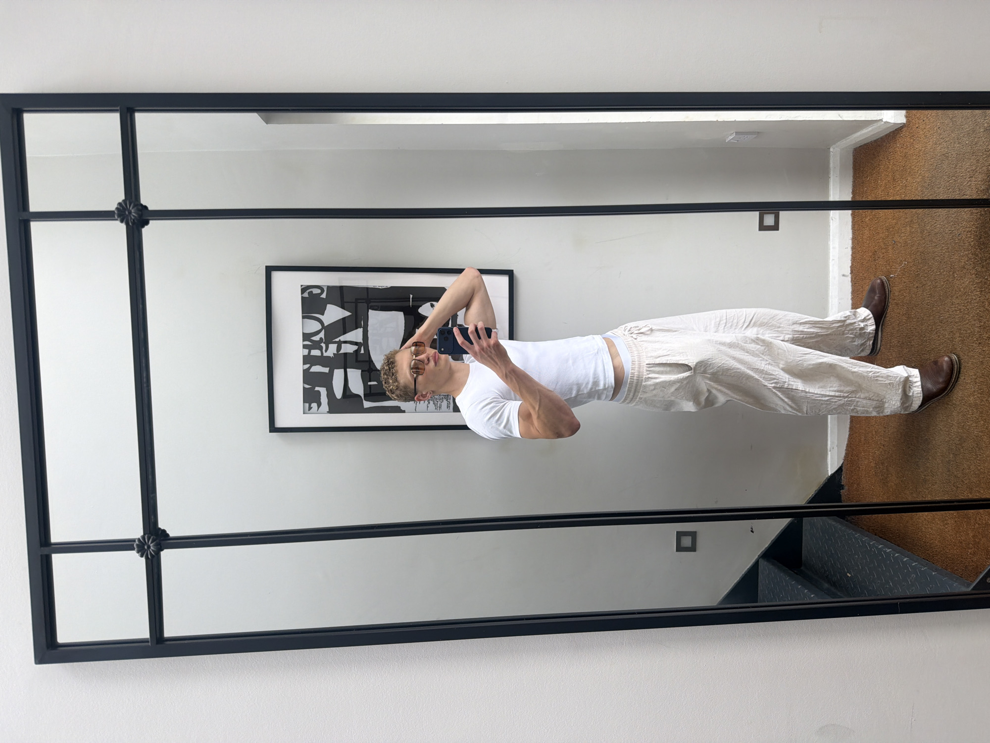 A man stands in front of a large, framed mirror, taking a selfie with his phone. He is wearing a white cropped t-shirt, loose-fitting light-colored pants, and brown shoes. He has short, curly blonde hair and is wearing sunglasses. The mirror is divided into panels by a black frame with decorative floral accents. In the reflection, a black and white abstract artwork hangs on the wall behind him. To the left of the artwork, a staircase with metal treads is visible. The room has white walls and a brown carpet. The image was taken in London, United Kingdom.