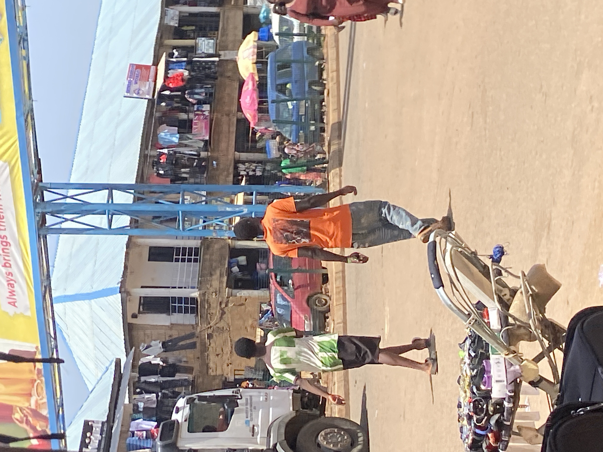 The image depicts a street scene in Bukuru, Nigeria. Two people are walking along a dusty road, passing a vendor's cart laden with goods. One person wears an orange t-shirt and jeans, while the other sports a white and green shirt with black shorts. Buildings and shops line the background, with clothing hanging on display outside. A white truck is parked on the side of the road.