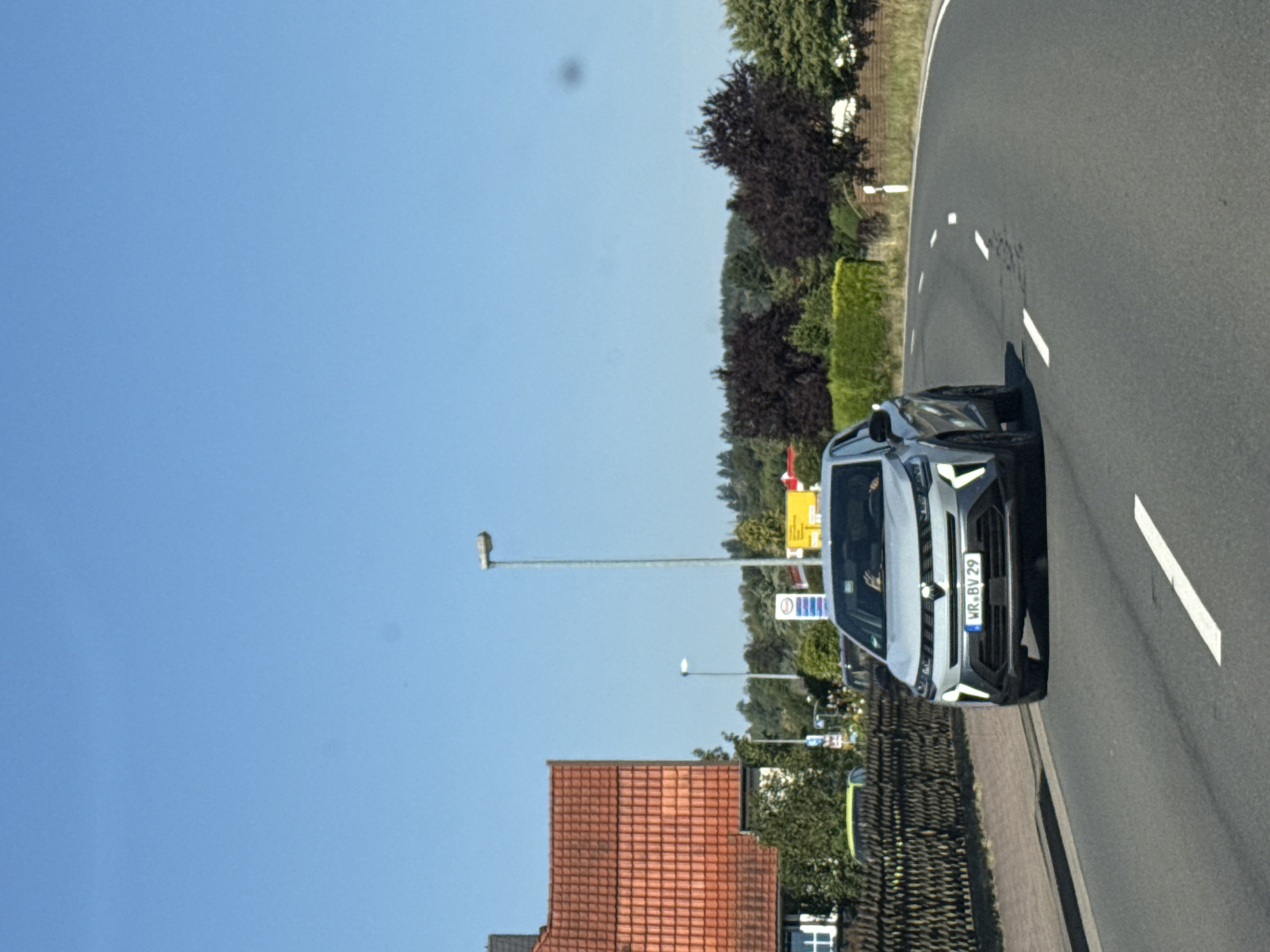 A light-colored SUV is driving on a paved road in Bad Sachsa, Germany. The SUV is in the foreground, and its license plate reads "WR.BV 29". The road curves to the right. To the left of the road, there is a sidewalk and a fence. Beyond the fence, there are houses and trees. In the background, there are more trees and hills. The sky is clear and blue. The sun is shining, casting shadows on the road.