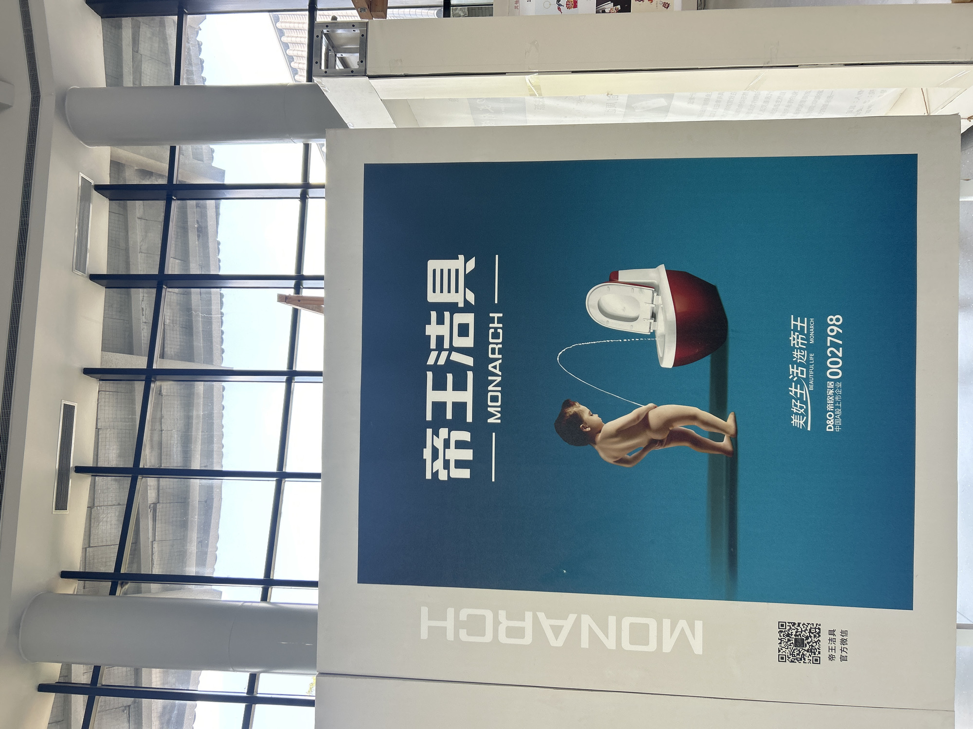 This image depicts a large advertisement for "Monarch" brand toilets, prominently displayed in what appears to be a public indoor space, possibly a mall or exhibition hall, in Jianyang City, China. The advertisement features a blue background and a visually striking image. In the foreground, a young boy, naked from the waist down, is shown urinating towards a red and white toilet. The stream of urine is depicted as a thin white line, curving and entering the toilet bowl. The brand name "MONARCH" is printed vertically in large white letters on the left side of the advertisement. Above the image of the boy and toilet, the Chinese characters "帝王洁具" (Dìwáng Jiéjù), meaning "Emperor Sanitary Ware," are displayed in large white font, followed by a smaller line that reads "MONARCH." At the bottom of the advertisement, there is a QR code, followed by Chinese text that translates to "Emperor Sanitary Ware Official WeChat." Beneath that, it says "Beautiful Life Select Emperor," with the English subtitle "BEAUTIFUL LIFE MONARCH." The D&O company name and a stock code are also visible. The background of the advertisement is a gradient of blue. The surrounding environment shows the upper parts of white cylindrical pillars and a grid-like ceiling structure with blue beams, indicative of a modern building's interior.