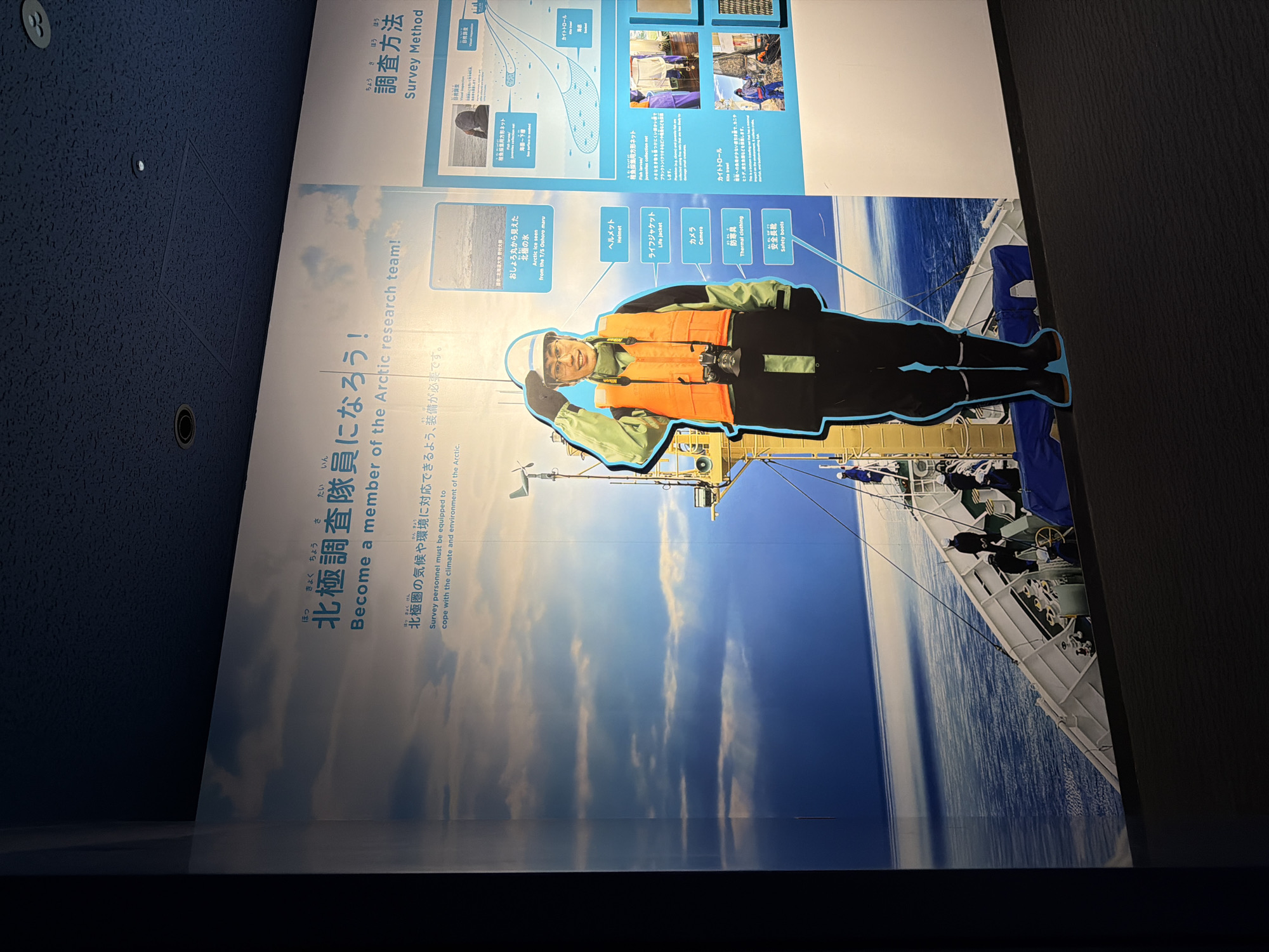 This image captures a brightly lit display wall in an indoor setting, likely a museum or exhibition, located in Osaka, Japan. The display's primary theme is Arctic research, inviting viewers to "Become a member of the Arctic research team!" and detailing "Survey Method."
The central visual features a life-sized cutout of a smiling person, presumably a researcher, dressed in appropriate Arctic gear: a white hard hat, an orange life jacket worn over a light green jacket, dark pants, and black boots. A camera is visible around their neck. This cutout is superimposed on a large background image of an open ocean with a research vessel under a partly cloudy, daylight sky.
Visible text on the display, primarily in Japanese with English translations, includes the titles "北極調査隊員になろう!" (Become a member of the Arctic research team!) and "調査方法" (Survey Method). Smaller panels list essential equipment: "ヘルメット" (Helmet), "ライフジャケット" (Life jacket), "カメラ" (Camera), "防寒具" (Thermal clothing), and "安全長靴" (Safety boots). Further details include descriptions and diagrams of research tools like "稚魚採集用方形ネット" (Fish larvae/juveniles collection net) and "カイトトロール" (Kite trawl). One caption references "おしょろ丸から見えた 北極の氷" (Arctic ice seen from the T/S Oshoro maru), indicating a specific research ship. The display aims to be educational, showcasing the practical aspects and equipment involved in Arctic exploration.