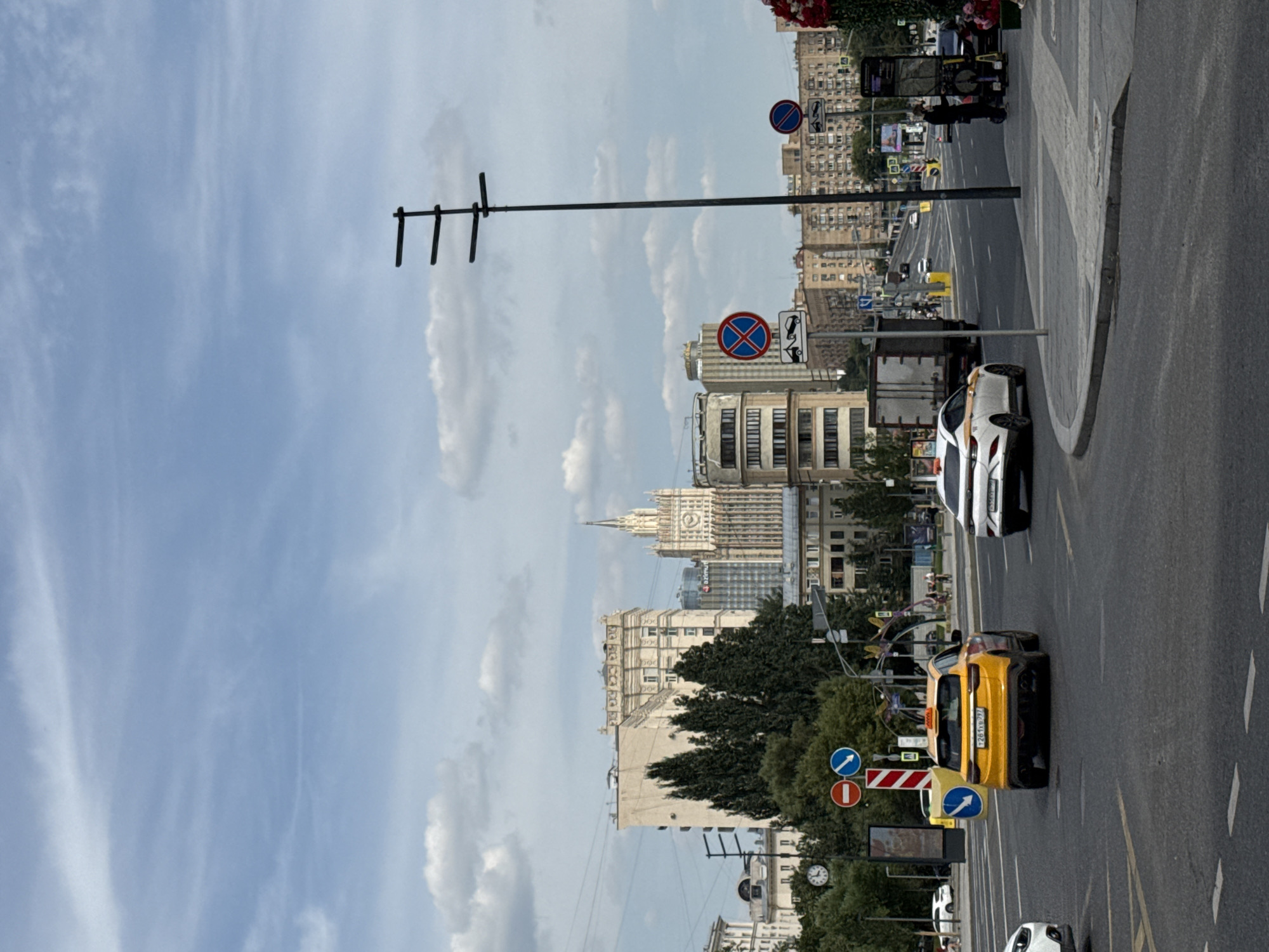This image depicts a street scene in Moscow, Russia, with a view towards several buildings, including a prominent Stalinist skyscraper. In the foreground, a yellow taxi is driving on a multi-lane road. To the left, traffic signs indicate no parking and a directional arrow. Further down the road, a white car and a truck are also visible. The sky is partly cloudy. In the background, various buildings of different architectural styles line the street, with the distinctive Stalinist skyscraper, likely the Ministry of Foreign Affairs building, standing out among them. The scene appears to be a typical urban environment with moderate traffic.