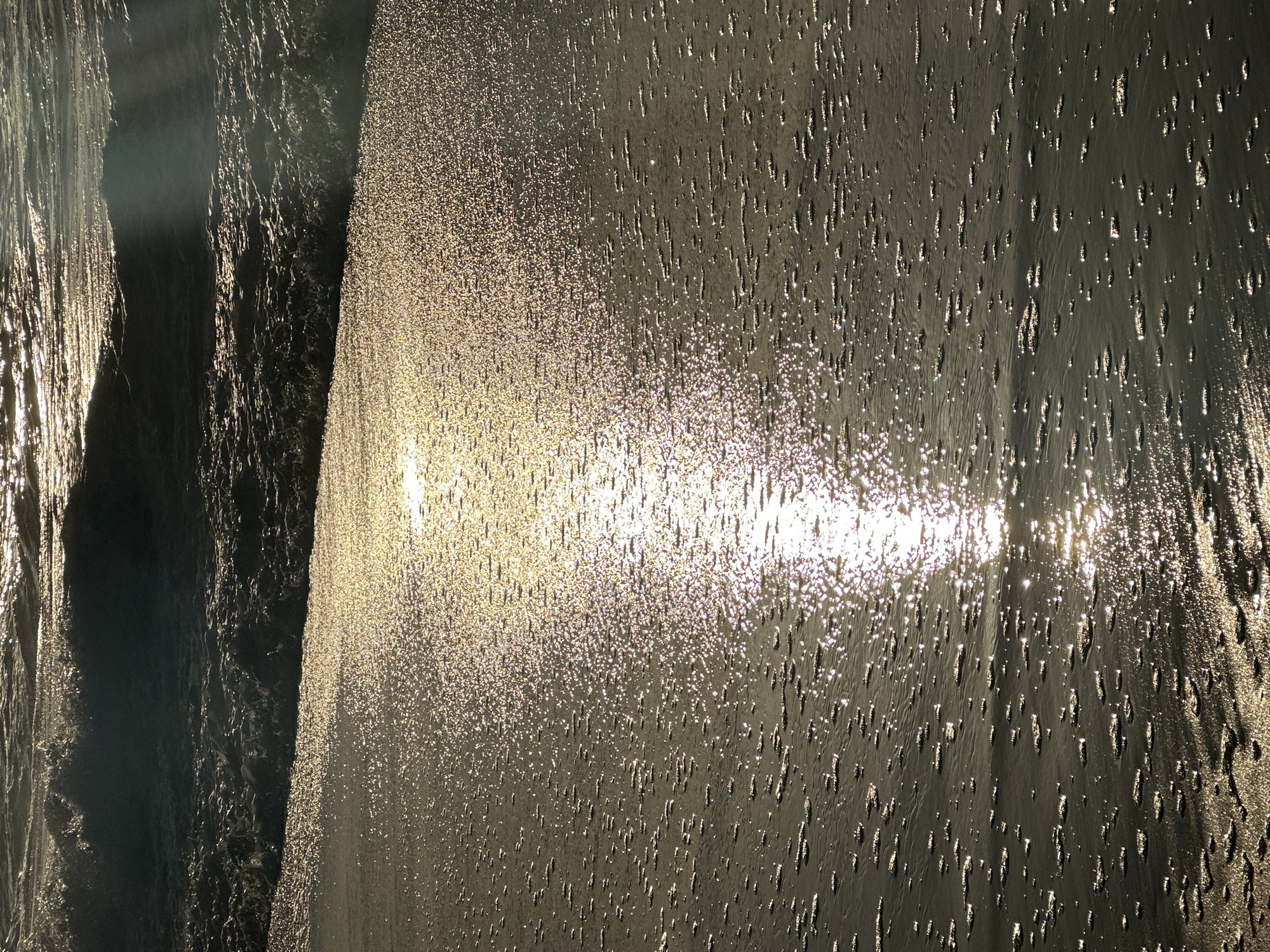 The image shows a close-up of the ocean at Mylestom, Australia, with the sun glinting off the water. The waves are breaking gently on the shore, and the sand is wet. The lighting suggests it is either sunrise or sunset. There are no people or other subjects visible in the image. The environment is a beach scene.
