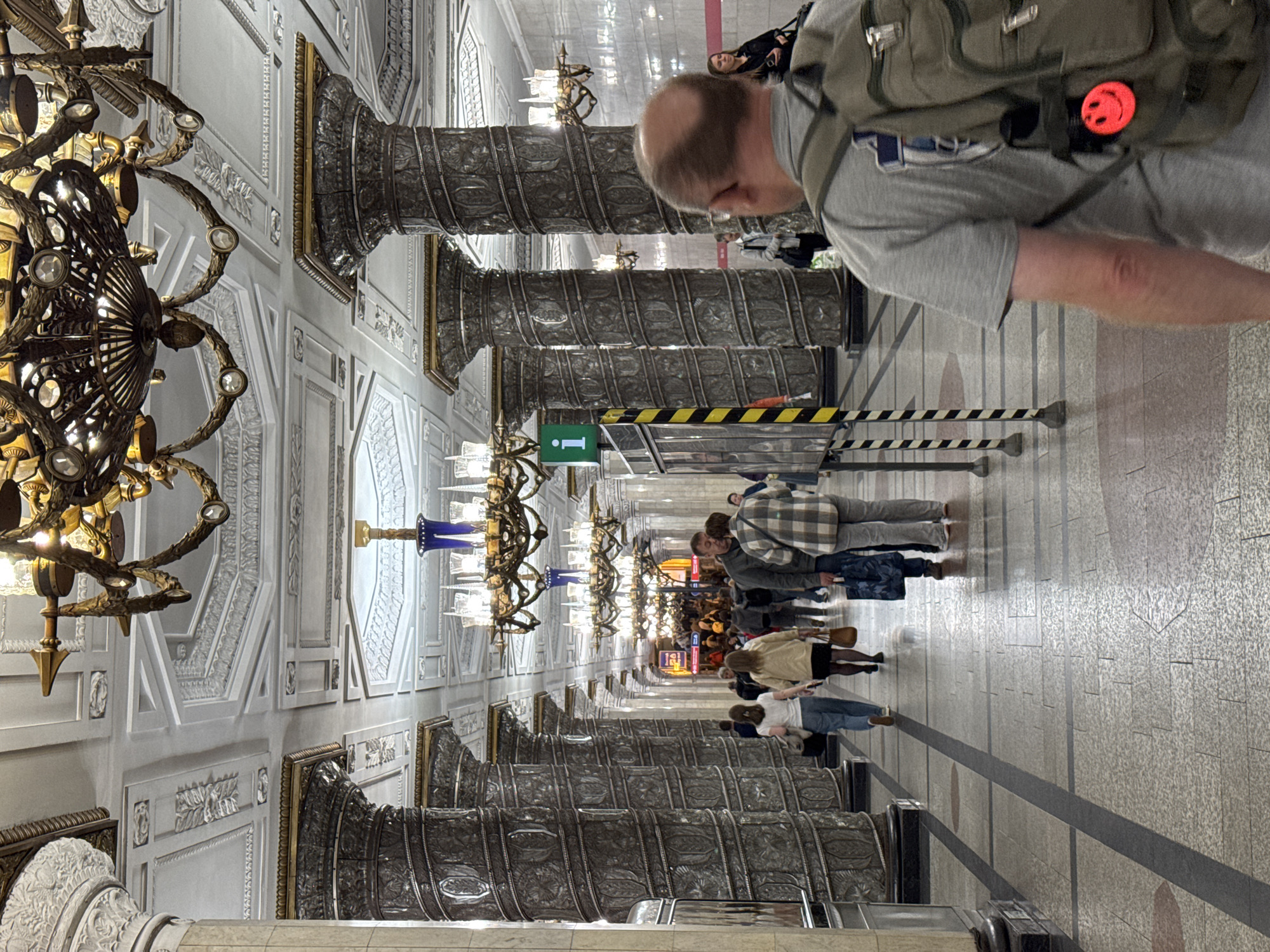 The image depicts the interior of the Komsomolskaya metro station in Saint Petersburg, Russia. The station is characterized by its ornate architecture, featuring massive, intricately carved columns that line the platform. Elaborate chandeliers hang from the high, decorative ceiling, and decorative lighting fixtures are mounted on the walls. Several people are visible walking along the platform. In the foreground, a man with a backpack is seen from behind. Further down the platform, a group of individuals are walking in various directions, some appearing to be tourists. The floor is tiled, with a pattern of dark and light grey tiles. A large information sign with a green "i" symbol is also visible on the platform. The overall impression is one of grandeur and historical significance, typical of many Saint Petersburg metro stations.
