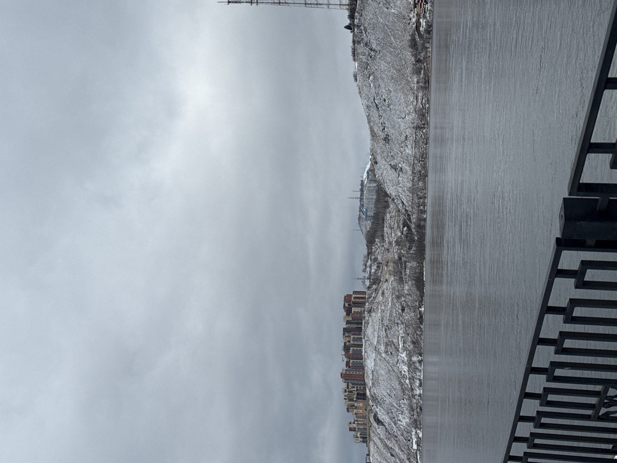 The image depicts a cityscape in Krasnoyarsk, Russia, taken on an overcast day. The sky is gray and filled with clouds. A series of apartment buildings are clustered along the edge of a snow-covered cliff or hill. A tall communication tower is visible at the top of the hill. A smooth, gray surface, possibly a walking path or overlook, is in the foreground. A section of a black metal railing is present, its design featuring a repeating geometric pattern.