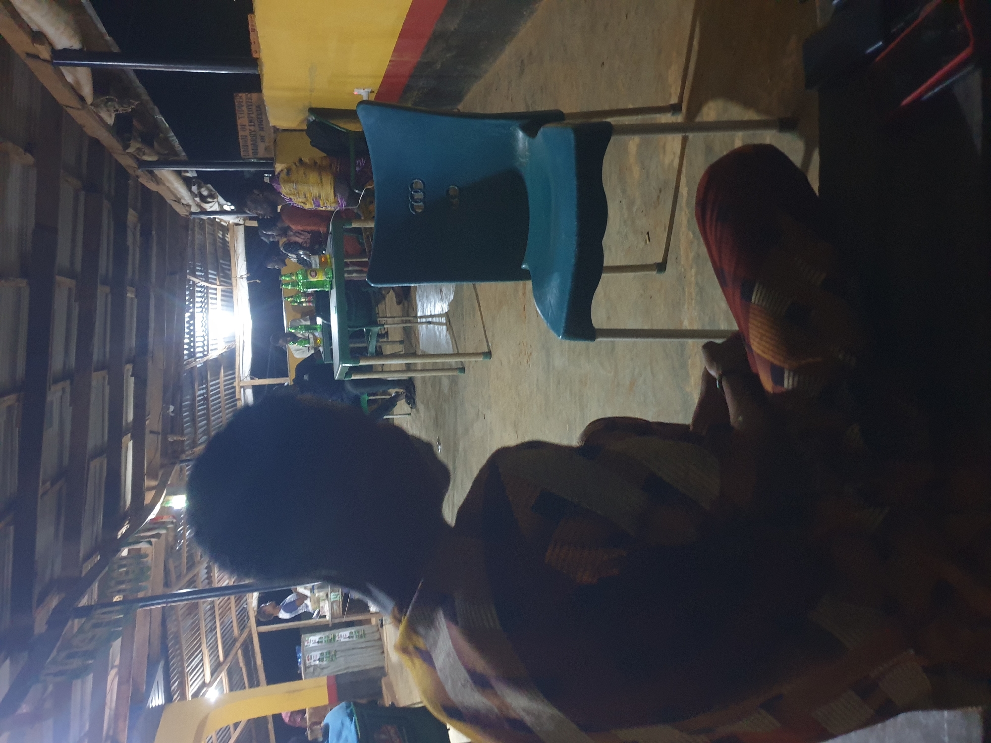The image depicts an outdoor seating area at night, likely a bar or restaurant, in Anilelerin, Nigeria. A person is seated in the foreground, facing away from the camera, wearing a colorful patterned garment. A bright blue plastic chair with silver legs stands empty in the middle ground, to the right. In the background, a group of people are seated at green tables. Several green beer bottles and glasses are visible on the tables. The seating area is covered by a corrugated metal roof supported by wooden beams. A yellow wall with a red stripe runs along the right side of the image. A sign is affixed to the yellow wall. The ground is a concrete surface. The lighting is dim, with overhead lights casting shadows.