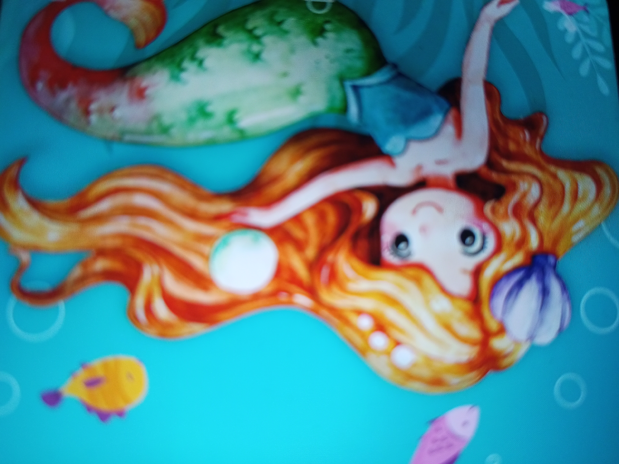 The image displays a vibrant, cartoon-style illustration of a mermaid floating underwater. The mermaid is positioned on her back, with her head towards the right side of the frame and her tail extending towards the left. She has long, flowing orange and red hair that spreads around her head. Her skin is light, and she wears a light blue top. Her tail transitions from a reddish hue at the fin to a mottled green on the main body. Her face shows a serene expression with large, dark eyes and a slight smile.
A purple seashell is nestled within her hair near her head, and a white pearl floats beside her extended left hand. Her right arm is raised gracefully upwards. The background is a clear, bright aqua blue, indicative of an underwater environment. Several faint circular bubbles are scattered across the blue background.
Two small fish are visible: a yellow fish with purple fins in the lower-left, and a pink fish in the lower-right, both swimming towards the left. Subtle green, leaf-like patterns, possibly seaweed, are visible in the upper right corner. The scene captures a whimsical and peaceful underwater moment. There is no visible text. This image was captured in Tinaquillo, Venezuela.