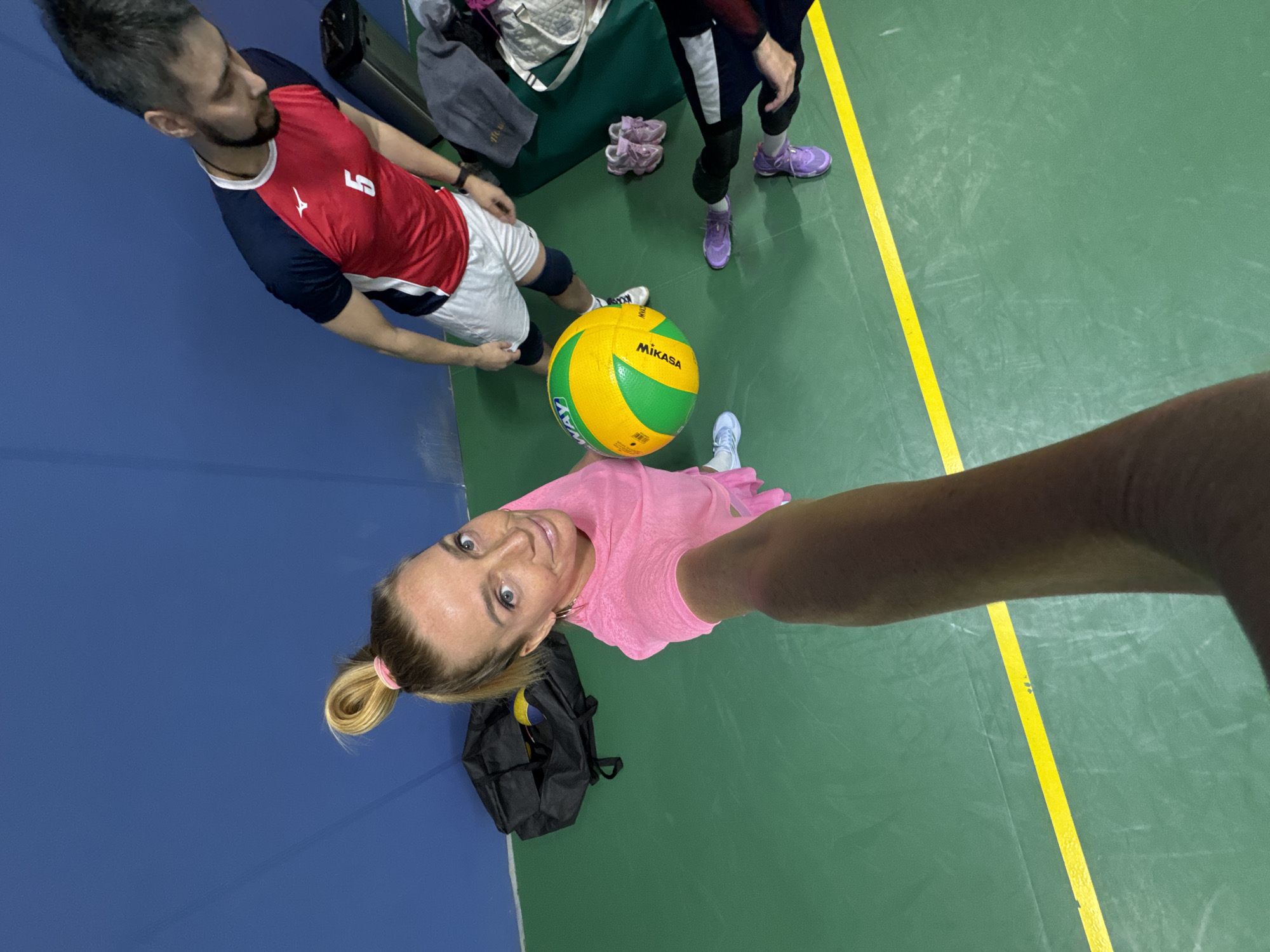 The image appears to be taken indoors, likely in a gym, with a woman taking a selfie. She is wearing a pink shirt and holding a Mikasa volleyball. To her left is a man wearing a red and navy blue jersey with the number "5" on it, and behind the volleyball, there is another person wearing purple sneakers. There's a dark blue wall on the left side of the frame and a green floor with a yellow stripe on the right. A black bag is on the floor near the woman, and what appears to be sports equipment and shoes are behind the man. There are no visual cues that suggest the location is in Saint Petersburg, Russia. The environment consists of elements commonly found in an indoor sports facility.