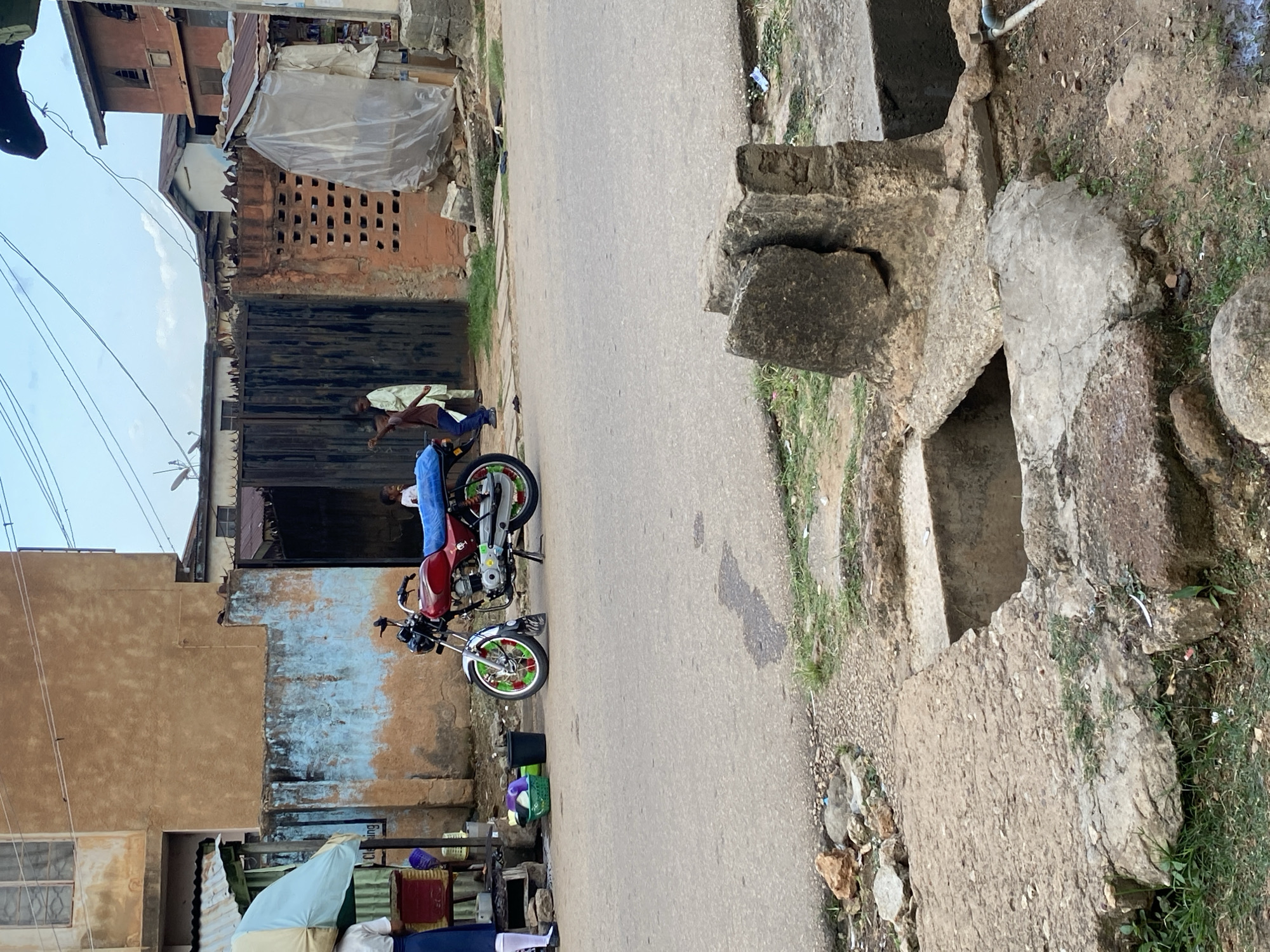 In a street scene in Jos, Nigeria, a red motorcycle is parked on the asphalt. The motorcycle has a blue cover on its seat and green and white accents on its wheels. To the left of the motorcycle, a young person in a white shirt and blue skirt stands near a stall with an umbrella, next to a green basket and a black bucket. In the background, there is a building with a textured, light brown facade and a section with peeling blue paint. A large, dark metal gate is partially visible, and beyond it, other buildings and structures can be seen. Two children are visible near the gate, one in a light-colored robe and the other in a dark shirt and pants, seemingly interacting. The street appears to be a public thoroughfare with some debris and vegetation on the side.