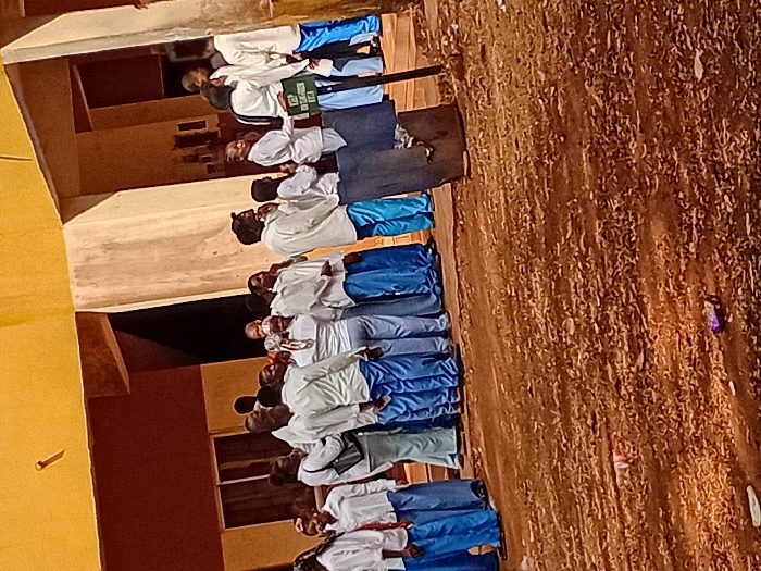 A group of people in school uniforms, consisting of white tops and blue bottoms, stand in a line, possibly for an event or assembly. They are positioned near a building with yellow and brown walls, with windows visible. The foreground shows a rough, dry ground covered in dirt and small debris. A sign is held by one of the individuals in the middle of the line. The scene has an outdoor setting with natural lighting.
