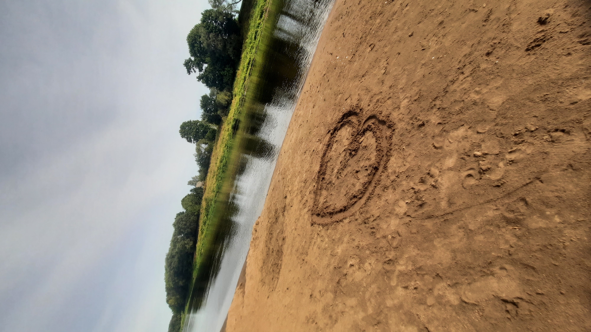 An outdoor, eye-level, slightly angled shot shows a wide river on the left, flowing towards the background, with a sandy embankment on the right. A heart shape has been drawn in the sand in the foreground. Lush green trees and vegetation line the opposite bank of the river, with a few houses visible amongst them. The sky is overcast. The location is the bank of the Dnieper River in the city of Shklov, Belarus.