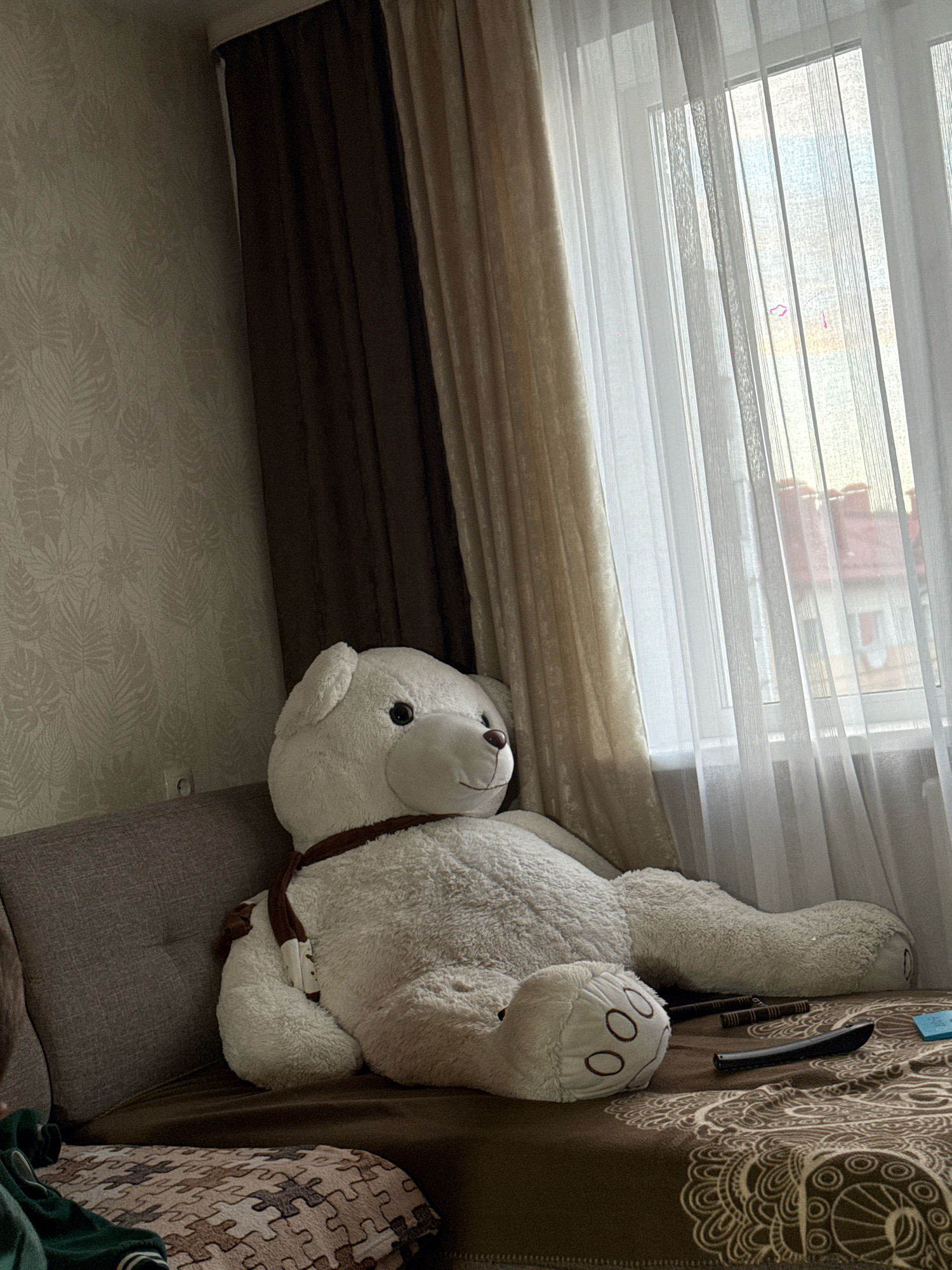 The image depicts the interior of a room, possibly a living room, with a large white teddy bear as the central subject. The teddy bear is seated on a brown sofa, positioned towards the left side of the frame. It is wearing a brown scarf around its neck. To the right of the teddy bear, sheer white curtains are drawn across a window, allowing natural light to enter the room. Outside the window, glimpses of buildings with red roofs are visible, suggesting an urban or suburban setting, potentially in the city of Slawharad, Belarus.
The sofa is covered with a brown throw blanket that has a patterned design. On the sofa, near the teddy bear's legs, there are two remote controls and a small light blue object, possibly a phone or a small box. A section of the wall on the left is visible, covered with textured wallpaper featuring a subtle leaf pattern. The overall lighting in the room is soft and natural, primarily coming from the window. The scene appears to be static, with no human activity or interaction evident.
