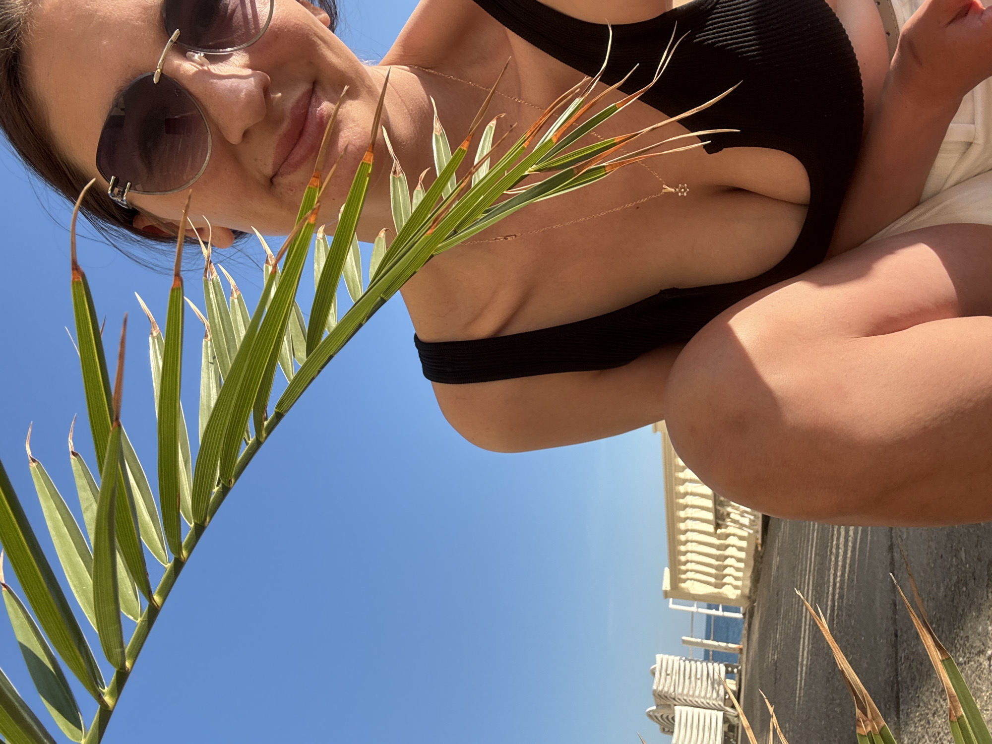 The image presents a low-angle perspective of a woman in sunglasses, posing outdoors. She is wearing a black top with a cutout and a light-colored bottom. A palm frond is held in the foreground, obscuring part of her face and torso. The background features a clear blue sky and a section of what appears to be a building or structure with repetitive vertical elements. Given the original data, it is likely that the location is Sochi, Russia.