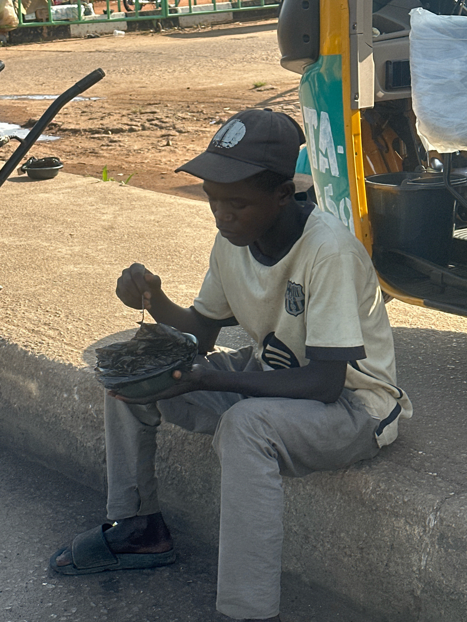 A young Black male wearing a dark baseball cap, a light-colored t-shirt with a dark collar and a crest on the left side of the chest, and gray sweatpants sits on a concrete curb. He is holding a dark bowl with black plastic wrapping inside and a small spoon or stick to retrieve items from it. He appears to be eating. His right foot is bare except for a dark sandal. Beside him on the curb is another dark bowl. Behind him is the side of a yellow and green auto-rickshaw. The ground is a mix of dirt and asphalt, with some loose gravel. The scene appears to be in Gura Top, Nigeria, possibly near a road or market.