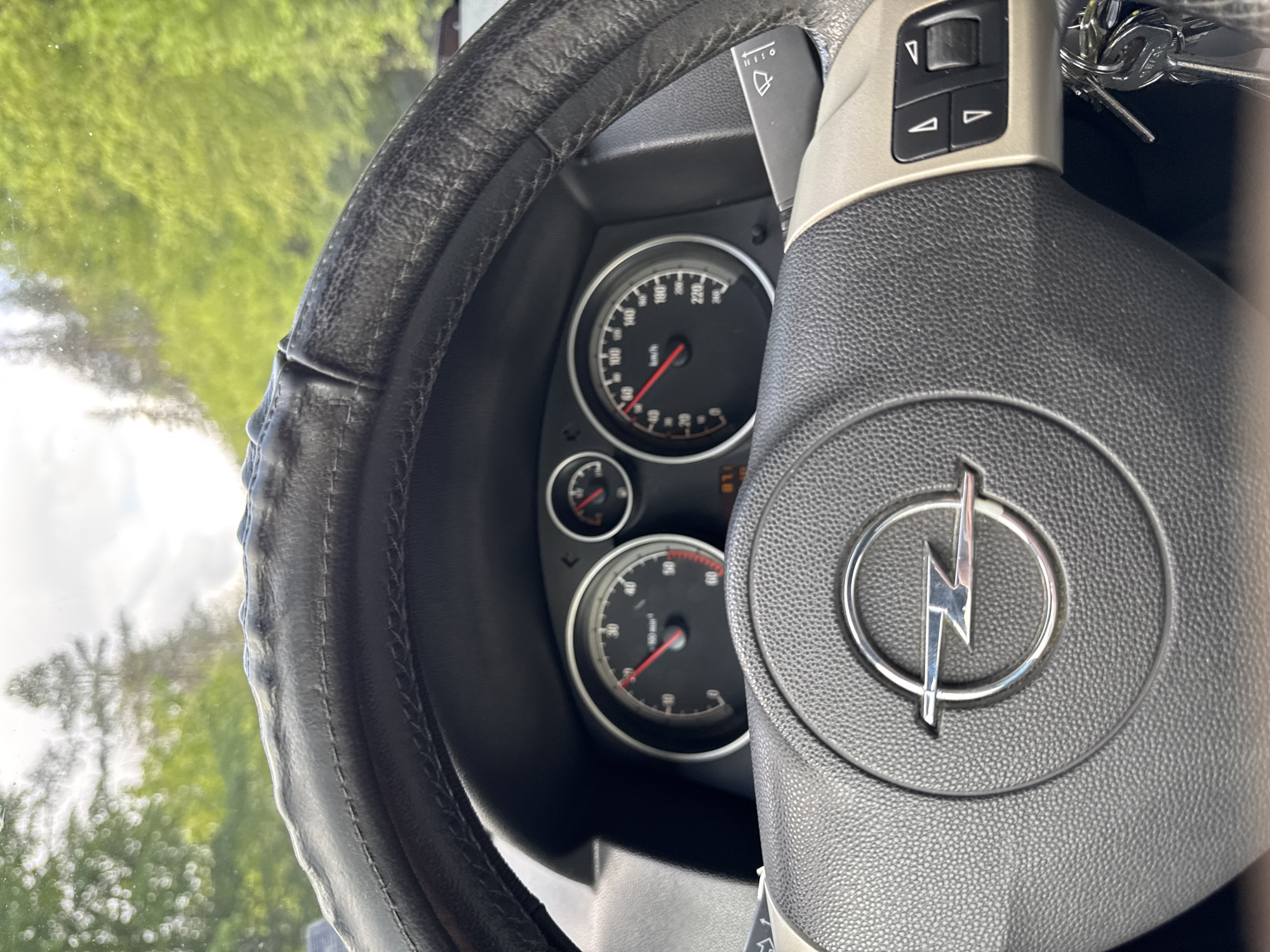 The image shows the interior of a car, specifically the steering wheel and dashboard. The steering wheel is black and has a worn leather cover. The Opel logo is prominently displayed in the center of the steering wheel. Behind the steering wheel, the car's instrument panel is visible, including the speedometer and tachometer. The speedometer indicates a speed of roughly 10km/h. Keys are visible in the ignition. The background is blurry but appears to show trees and foliage. There is no information in the image to associate the location to Chokmanovo, Bulgaria.