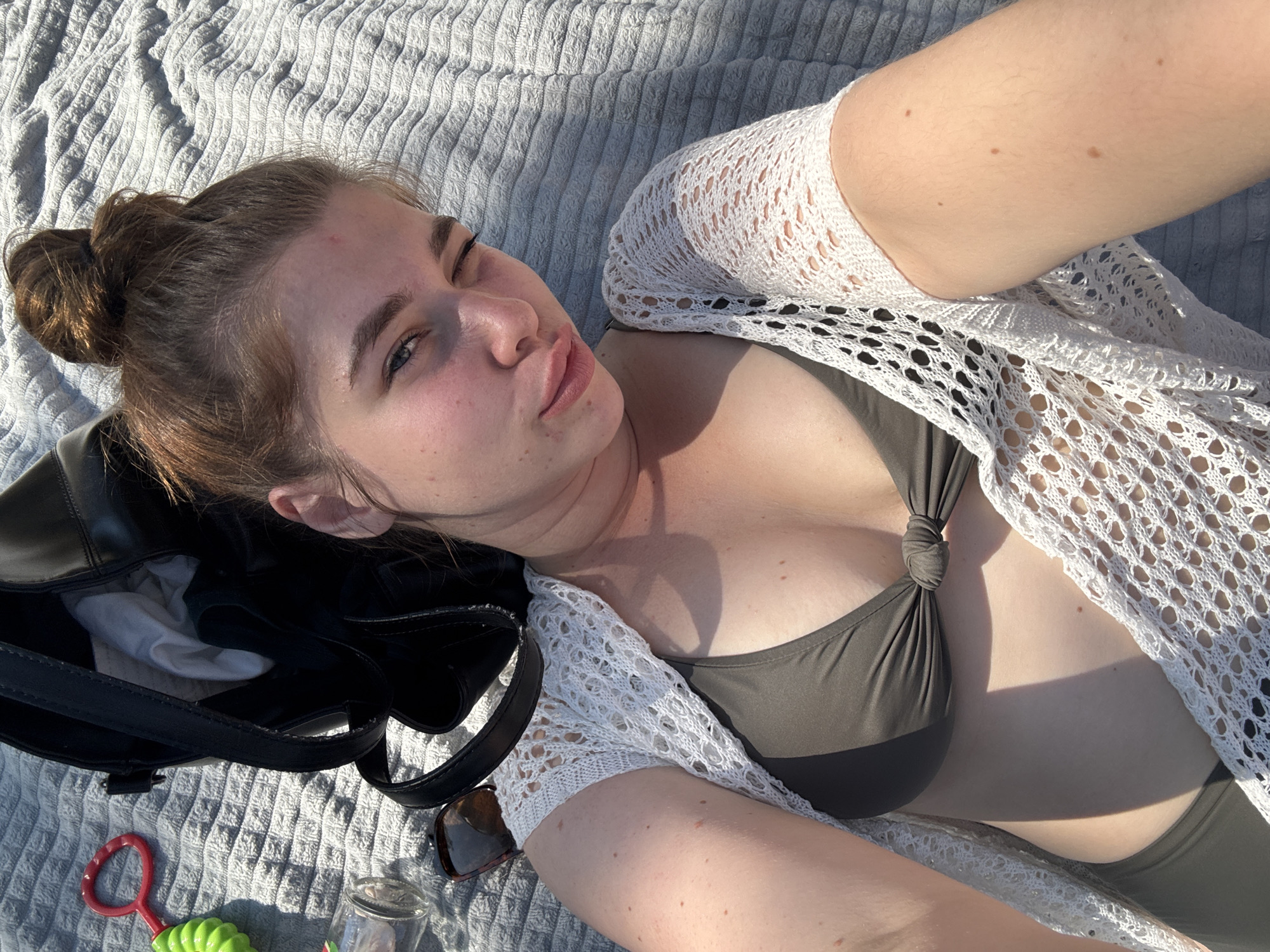 This is a close-up, high-angle selfie of a young woman lying on a textured gray blanket outdoors. The woman has her hair pulled up in a bun and is wearing a dark green bikini top and a white, lacy cover-up. She is looking towards the camera with one eye squinted and her lips pursed. Her skin appears flushed, suggesting she may be enjoying the sun.
To her left, a black bag is visible, with some white fabric peeking out. Near the bag, a pair of sunglasses and a small glass jar are placed on the blanket. In the lower left corner, a child's toy, a red ring attached to a green handle, is also present.
The lighting suggests it is daytime, with sunlight casting shadows, indicating a sunny day. The background consists solely of the textured blanket, offering no further clues about the specific location other than it being outdoors. The overall mood appears relaxed and casual. There is no visible text in the image.
