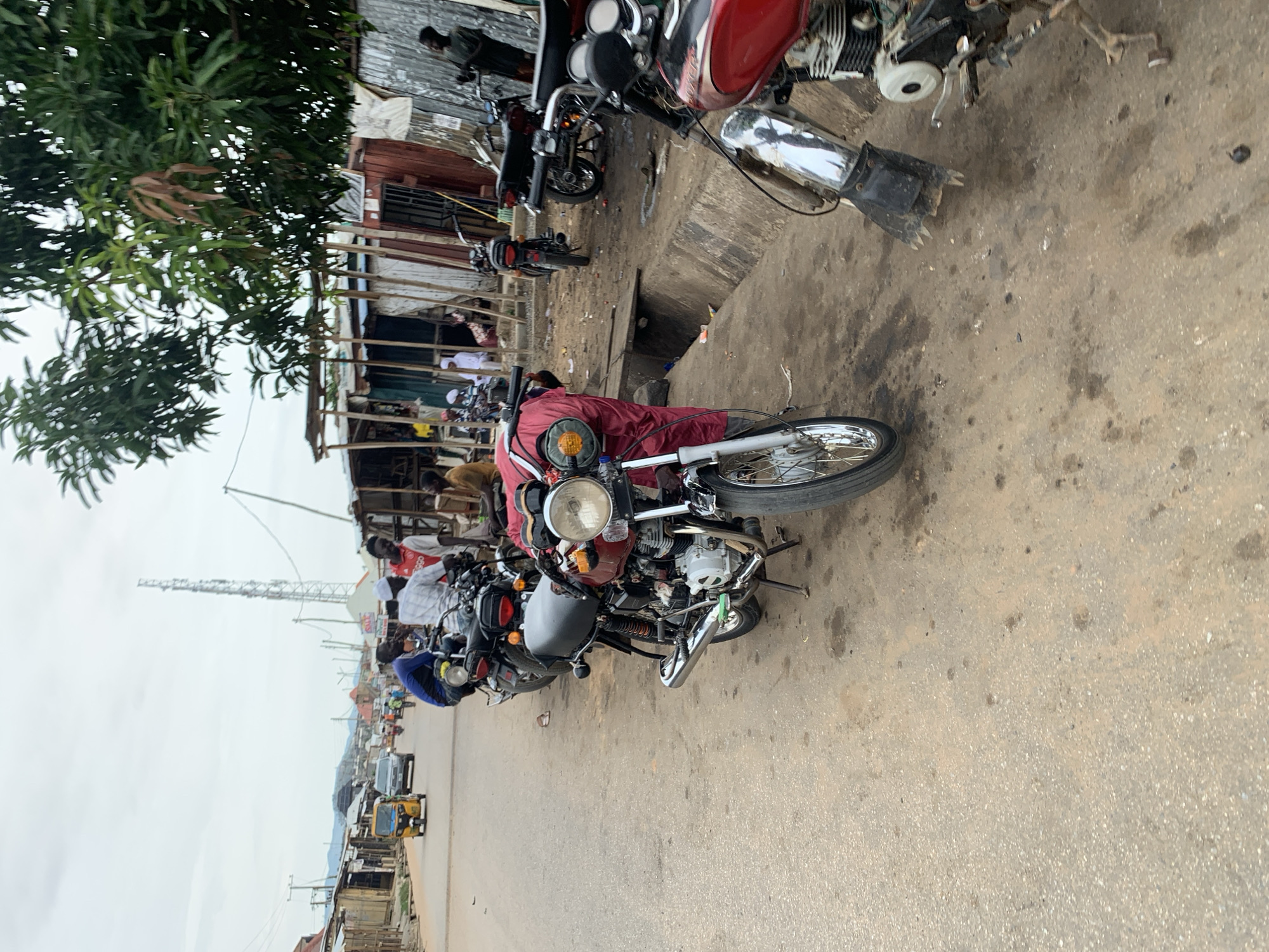 This is a street-level, medium-shot, outdoor photograph taken in Abuja, Nigeria. The scene depicts a roadside with several motorcycles parked. In the foreground, a red motorcycle is prominently featured, its chrome handlebars and silver spokes gleaming. Behind it, a line of other motorcycles, primarily dark-colored, are parked along the dirt and asphalt road. A red cloth is draped over the back of the first motorcycle. Several people are visible in the background, some standing near the motorcycles and others appearing to be engaged in activities in the market stalls that line the street. The market stalls are constructed from corrugated metal and wood, suggesting a local commercial area. A yellow tricycle, commonly known as an "Okada" or "Keke," is visible further down the road, indicating traffic and transportation in the area. The sky is overcast, suggesting a cloudy day. A tall communication tower is visible in the distance, a common feature in urban landscapes. The overall impression is of a busy, informal marketplace in a Nigerian city.