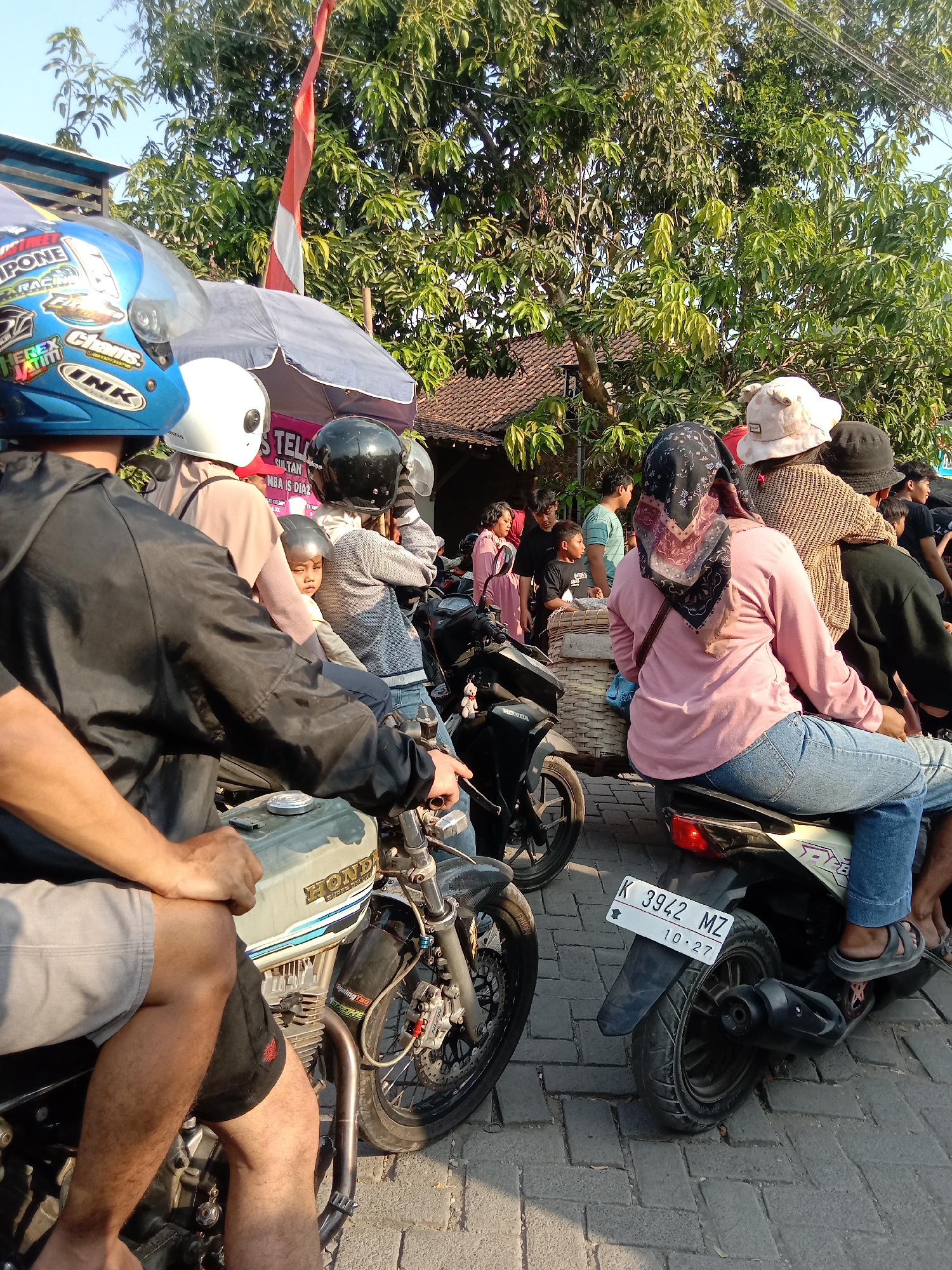 The image depicts a street scene in Wirosari, Indonesia, likely during daytime hours. A group of people are gathered, many of them on motorcycles, suggesting a market or public event. In the foreground, a person wearing a blue helmet and a black jacket is seated on a motorcycle, with their hands resting on the handlebars. Beside them, another person in a white helmet is also on a motorcycle, and behind them, a child in a grey helmet is visible. Further back, several other individuals are on motorcycles and standing, some carrying bags or baskets. The environment includes trees, buildings with tiled roofs, and a red and white Indonesian flag fluttering in the background. The road appears to be paved with interlocking stones. A license plate on one of the motorcycles reads "K 3942 MZ 10.27". The overall impression is one of a bustling, everyday scene in a busy Indonesian town.