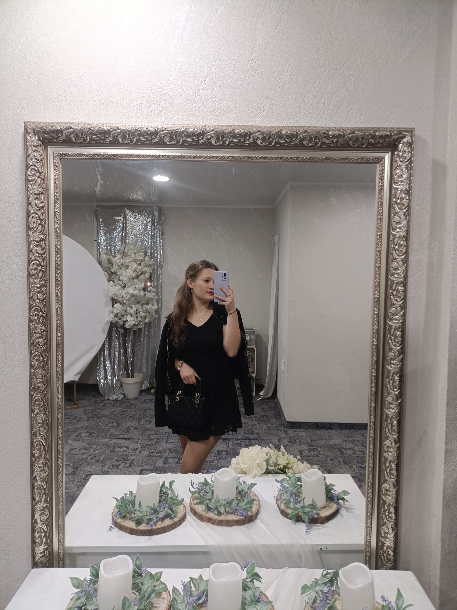 A woman is taking a selfie in a mirror. The mirror is large with an ornate silver frame. In the reflection, the woman is wearing a black dress and has a black jacket over her shoulder. She's holding a mobile phone in her right hand. Behind her is a decorated event space with a silver sequin backdrop, a white blossom tree in a pot, and a white circular screen. In front of the mirror, a table is decorated with white fabric, faux flower arrangements, and candles on wooden bases. The location appears to be an indoor event space, potentially in Plovdiv, Bulgaria, given the prompt's suggestion, although there are no distinctive features to definitively confirm this.