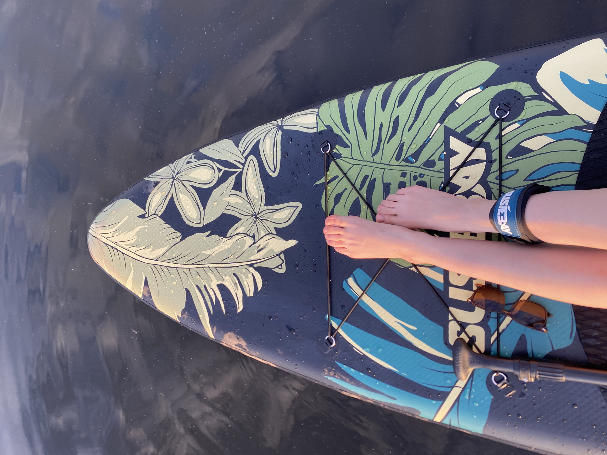 The image shows a person sitting on a paddleboard on water. The paddleboard has a design with green and blue tropical leaves and flowers on a dark background. There are water droplets on the paddleboard's surface. The person's bare feet are visible towards the front of the board. The person is also wearing a blue wristband on their arm. A pair of sunglasses and part of a paddle are resting on the paddleboard. The water is dark with subtle reflections. There is no indication in the image to support a location within the city of Chaikovsky, Russia.