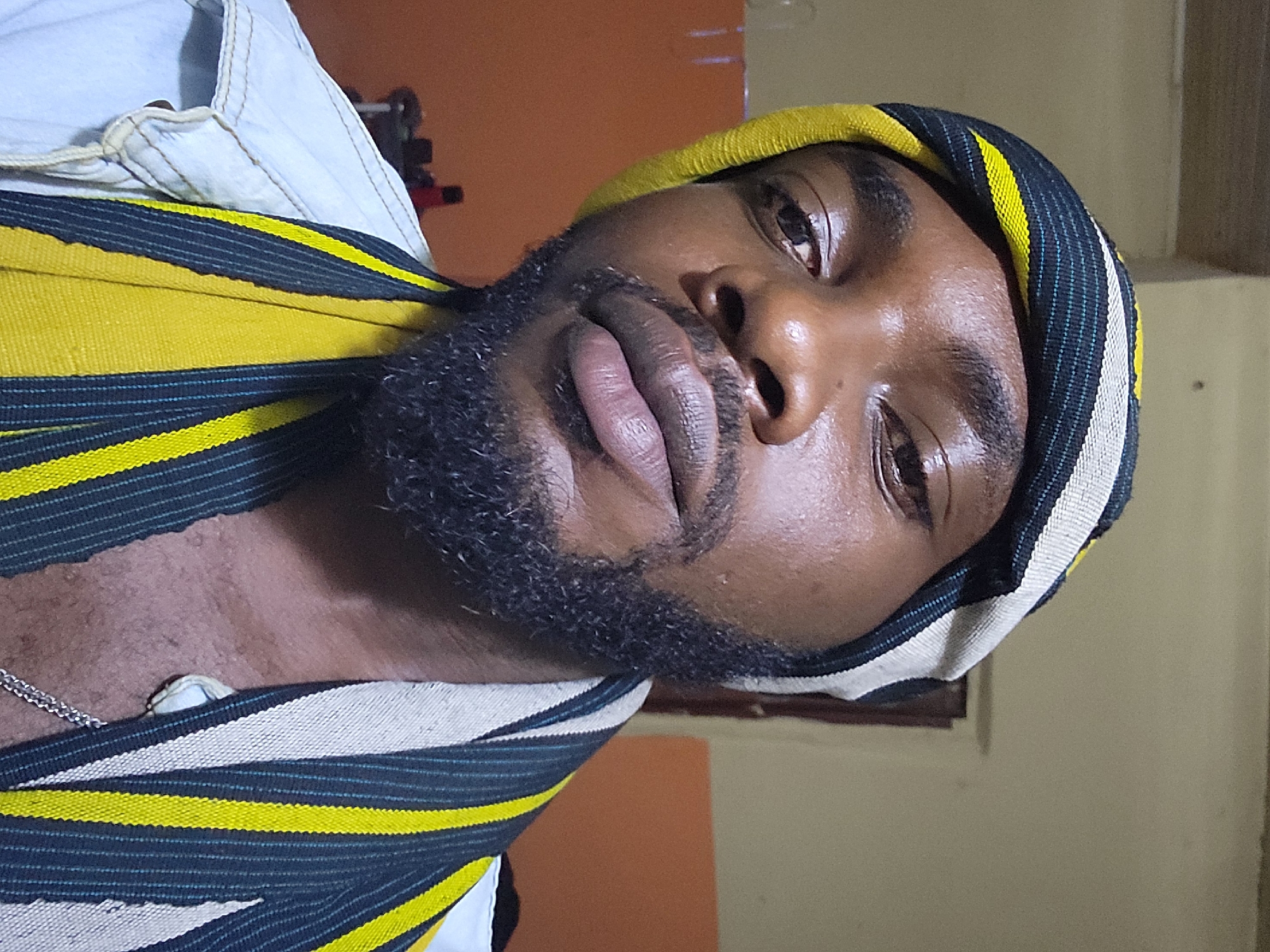 A close-up, indoor portrait shows a dark-skinned man with a beard. He is wearing a woven cloth head covering and a white shirt. A silver chain is visible at his neck. The location is possibly Jos, Nigeria, based on the man's appearance. The background is blurry and suggests an indoor setting. No other details are discernible from the image.