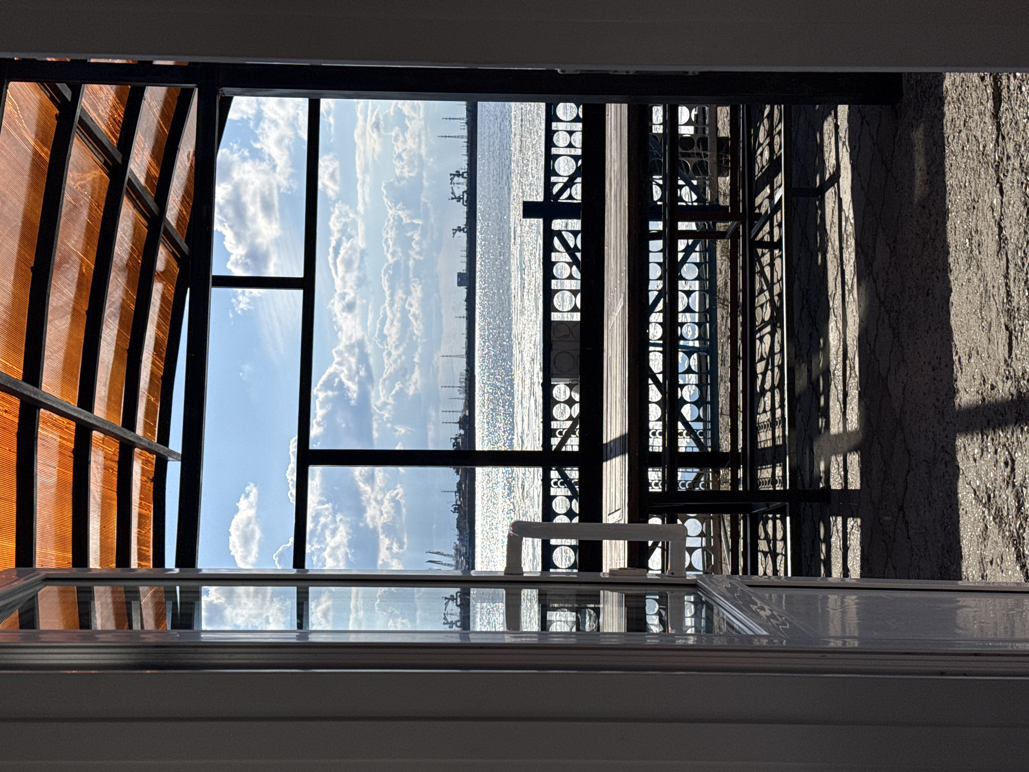 The image, when viewed with a horizontal orientation, presents a serene daytime scene looking out from an enclosed or semi-enclosed structure over a large body of water in Chaikovsky, Russia.
In the upper-left, a translucent, corrugated canopy of an orange-brown hue provides shade, supported by dark metal beams, suggesting a patio or veranda roof. Adjacent to it, a clear blue sky, adorned with scattered white cumulus clouds, dominates the upper part of the frame. Below the sky, a vast expanse of water stretches across the middle of the image. The water surface intensely reflects the bright sunlight, creating a shimmering, sparkling effect. Along the distant horizon, faint silhouettes of industrial structures, possibly cranes or factory buildings, are visible against the bright sky, hinting at a port or industrial area along the Kama River or Votkinsk Reservoir.
To the right of the water, an intricate dark metal structure, characterized by numerous circular cutouts, acts as a barrier or decorative architectural element. Further to the right, a textured, dark grey wall, likely concrete or stone, is visible, marked by strong shadows. In the immediate foreground, at the bottom of the image, a white window frame indicates the vantage point, reflecting parts of the scene above. A white handle is also visible on the window frame.
The scene captures a moment of quiet observation. The bright sunlight, clear sky, and sparkling water suggest fair weather during the day, likely morning or afternoon. No people or specific activities are depicted. There is no visible text in the image.