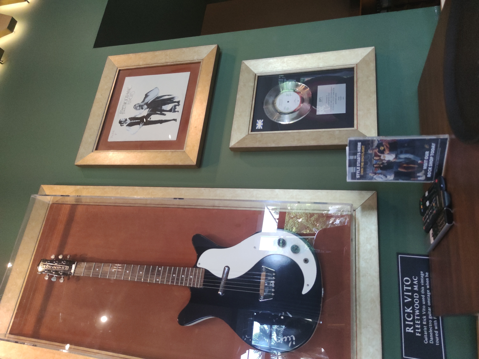The image depicts a wall in what appears to be a venue in Podgorica, Montenegro. Displayed on the green wall are several framed items related to the band Fleetwood Mac. At the top left, a vintage black and white electric guitar with a yin-yang-like white pickguard is housed in a glass case. Below the guitar, a black plaque reads "RICK VITO FLEETWOOD MAC Guitarist Rick Vito used this vintage Danelectro guitar onstage when he toured with F." To the right of the guitar, a framed album cover for Fleetwood Mac's "Rumours" is displayed. Beneath that, a framed platinum record award, featuring a silver disc and a plaque with text and a Union Jack, is mounted. In the foreground, on a wooden surface, are several remote controls and a small promotional stand with an image of people and text that reads "STYLE STARTS HERE OWN THE SPOTLIGHT VISIT OUR ROCK SHOP TODAY." The lighting suggests the scene is indoors, with spotlights visible at the top of the image.