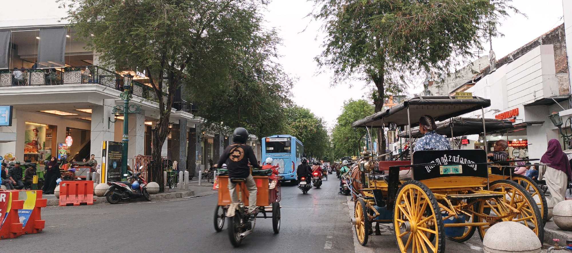 The image depicts a street scene in Yogyakarta, Indonesia, likely Malioboro Street, a well-known tourist destination. The street is bustling with activity, featuring a mix of traditional and modern transportation. In the foreground, a traditional horse-drawn carriage, known as a "andong," is prominently displayed with its yellow wheels and ornate decorations. The carriage is occupied by at least two people, one of whom appears to be a driver wearing a traditional Javanese headwrap.
To the left of the carriage, several motorcycles and a three-wheeled motorized vehicle are visible, indicating the presence of more contemporary transport. A blue bus is also seen further down the street, suggesting public transportation is available. The street is lined with buildings, some appearing to be shops or restaurants, with awnings and balconies characteristic of urban Indonesian architecture. Green trees with dense foliage line the street, providing shade and a natural element to the cityscape.
Several individuals are present in the scene, including pedestrians dressed in various attire, some wearing headscarves. One person on a motorized tricycle is wearing a black long-sleeved shirt with a circular graphic and a helmet. The overall atmosphere suggests a busy commercial and tourist area, capturing the vibrant daily life of Yogyakarta. The lighting indicates it is daytime, possibly late afternoon or early morning due to the somewhat overcast sky.