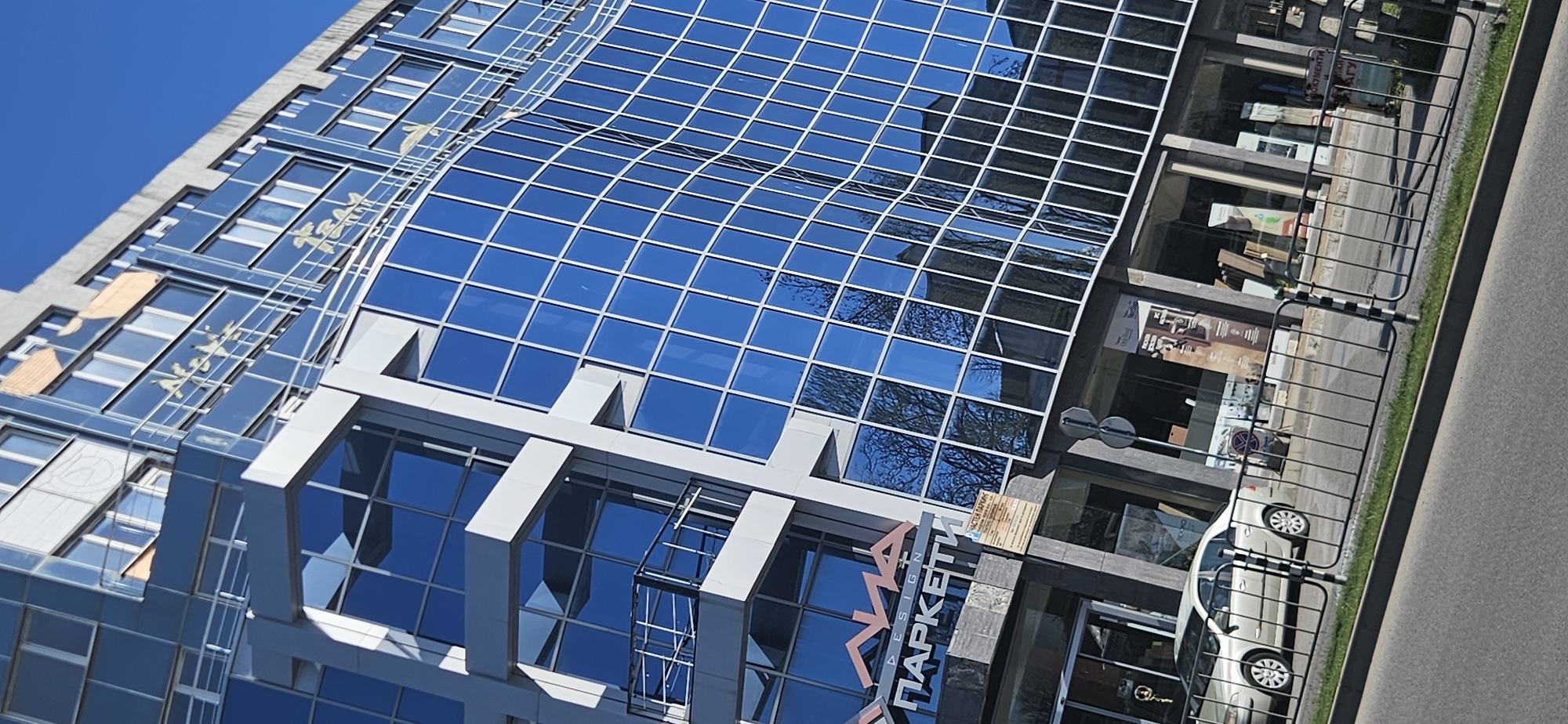 The image shows a modern glass-facade building in Plovdiv, Bulgaria. The building has a curved glass section and a sign that reads "DESIGN ПАРКЕТИ" in Cyrillic. Reflections of trees and the sky are visible on the glass. A sidewalk, road, and metal railing are in the foreground. A white car is parked near the building.