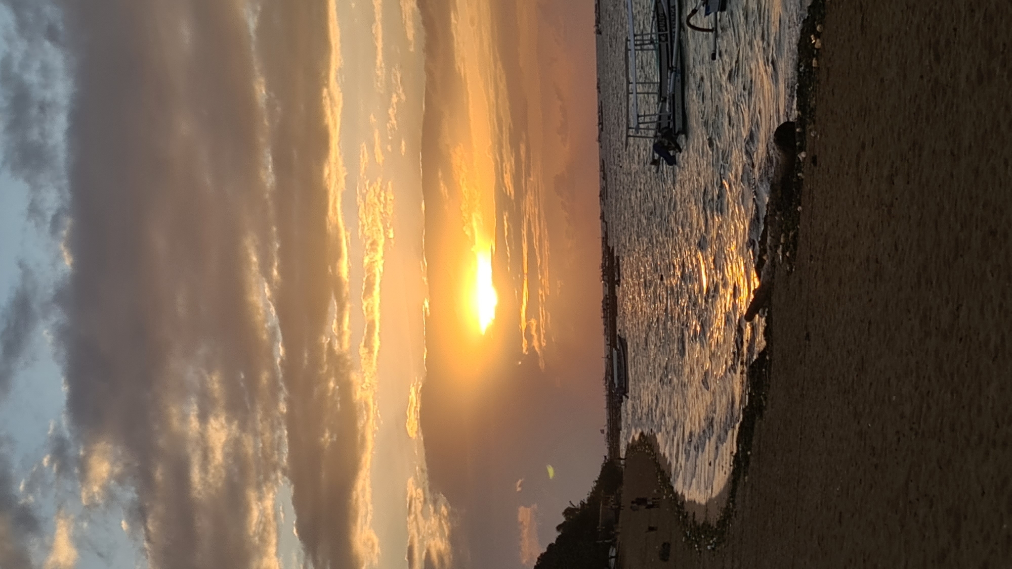 The image depicts a sunset scene at a beach, likely in Denpasar, Indonesia, given the context. The sky is filled with dramatic clouds tinged with orange and yellow from the setting sun, which is partially obscured by clouds and casting a bright glow across the water. The sea is calm with gentle waves lapping against the shore. Several small boats are visible anchored in the water. The beach itself is sandy and appears to have some debris scattered along the shoreline. A line of trees can be seen in the distance on the left side of the image, and a few people are walking along the beach. The overall impression is one of a serene and picturesque coastal evening.