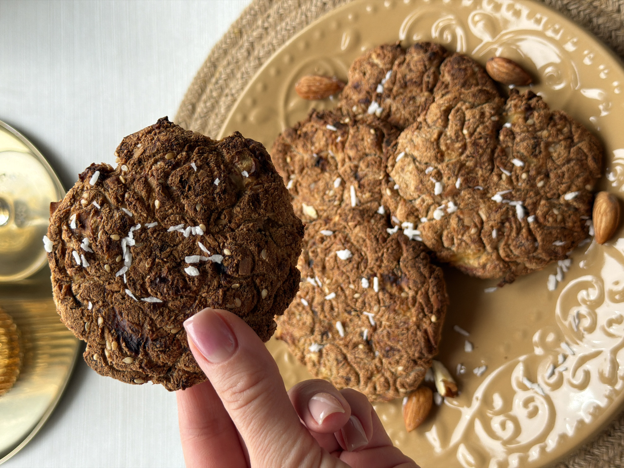 A hand is holding a brown, textured cookie with scattered white flakes. The cookie's surface has a slightly rough texture. In the background, there are three more similar cookies on a light brown plate decorated with almonds. The plate sits on a round, woven placemat. To the left, a gold-colored object is partially visible. The background surface appears to be a white, textured material. The location cannot be determined from the image.