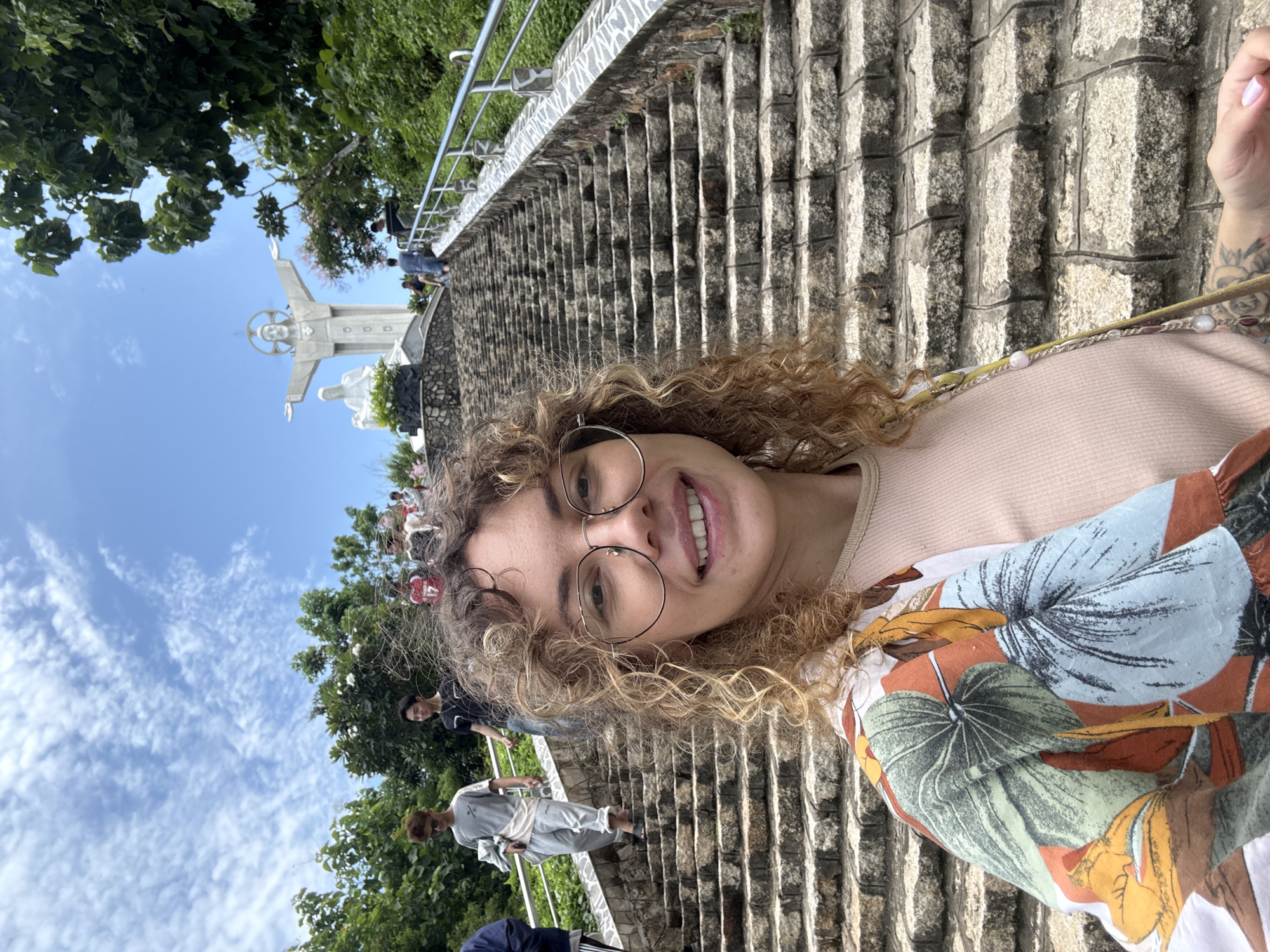 The image shows a young woman with curly hair and glasses taking a selfie on a stone staircase. Behind her, a statue of Jesus Christ with outstretched arms is visible at the top of the stairs, along with other people walking up the stairs. The scene appears to be a tourist attraction, possibly a religious site or a viewpoint. The sky is blue with white clouds, and there is lush green vegetation surrounding the area. The woman is wearing a light pink top and a colorful patterned shirt. This location is likely the Christ the King statue on Ba Den Mountain in Tay Ninh province, near Ho Chi Minh City, Vietnam, which is a popular pilgrimage site and tourist destination.