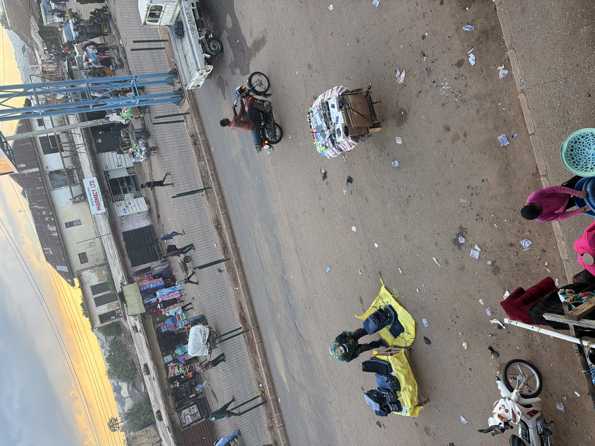 The image depicts a street scene in Bukuru, Nigeria. The sky is overcast with a hint of golden light suggesting it might be early morning or late afternoon. In the background, a collection of buildings with corrugated iron roofs line the street. Some of these appear to be shops, with displays of clothing visible in one establishment.
The foreground of the image is dominated by a paved road and sidewalk area, with a dusty, unpaved section to the left. Several people are present, engaged in various activities. A man in a red and black striped shirt is riding a motorcycle down the road. Another man, wearing a green camouflage jacket, is standing near a yellow tarp displaying stacks of blue jeans. A cart piled high with various items, possibly electronics, is being pushed by an unseen person. Further down the road, a white pickup truck is parked.
On the sidewalk area, a person wearing a pink long-sleeved shirt is interacting with blue and green plastic containers. Another individual in a dark garment is partially visible behind a wooden structure. The ground is littered with various debris, including plastic bags, wrappers, and what appear to be crumpled pieces of paper. A white motorcycle is parked on the unpaved section of the ground. The overall scene suggests a busy market or commercial area, with a mix of vehicular and pedestrian traffic and street vendors.