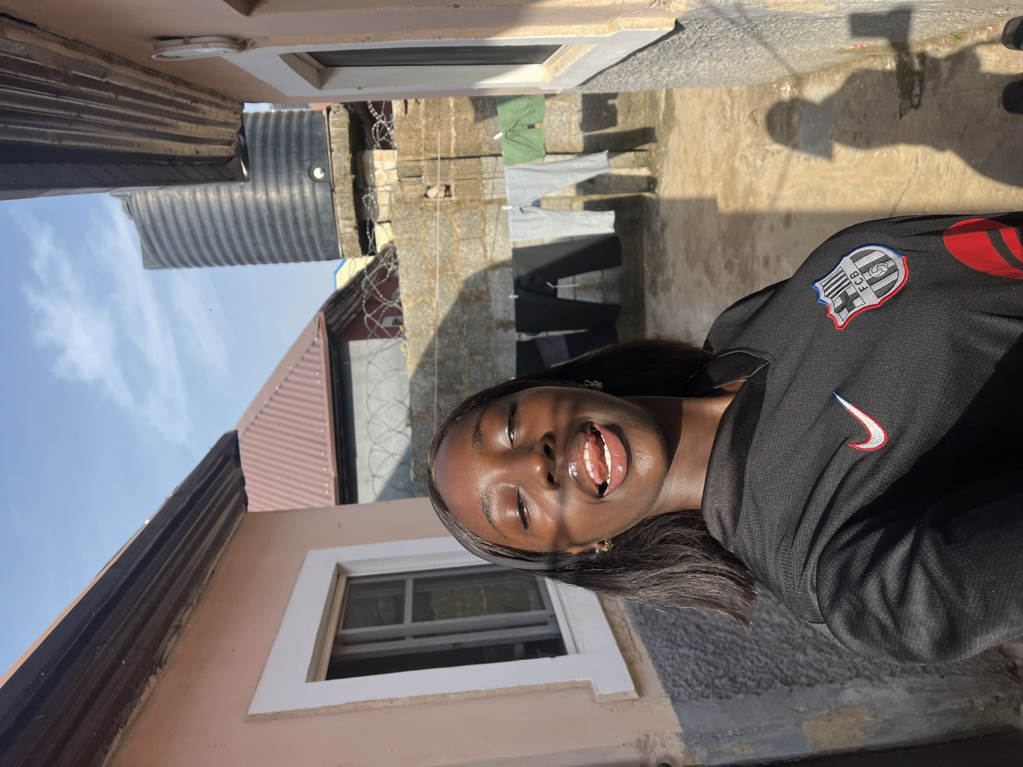 A young, dark-skinned woman smiles broadly, mouth open, in an outdoor setting. She wears a black jacket with a Nike logo and a crest featuring "FCB". The surroundings include a building with a window and a pinkish-beige exterior, a corrugated metal roof, and a clear blue sky. Clothes are hanging on a clothesline strung between buildings in the background. The scene is likely in a residential area, potentially in Abuja, Nigeria, based on context provided.
