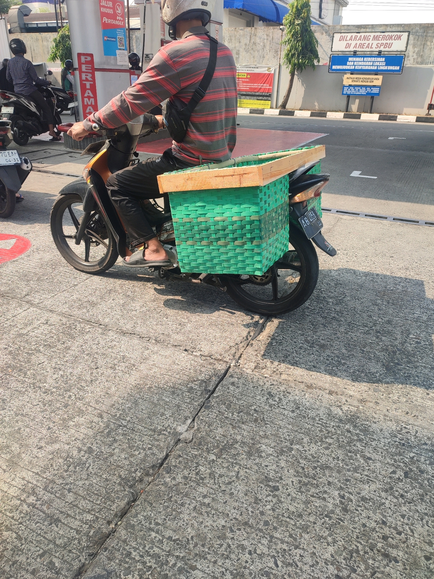 This is a medium shot, taken from a slightly elevated perspective, of a man riding a motorcycle in Yogyakarta, Indonesia. The motorcycle is black and orange and has a large green woven basket attached to the rear, which appears to be carrying something with a wooden frame. The man is wearing a helmet and a long-sleeved striped shirt, black pants, and sandals. He is looking forward. The background shows a Pertamina gas station with a sign that says "PERTAMAX". There are other motorcycles and people in the background. The ground is paved with concrete. The lighting suggests it is a sunny day. The image appears to have been taken at street level, possibly from another moving vehicle.