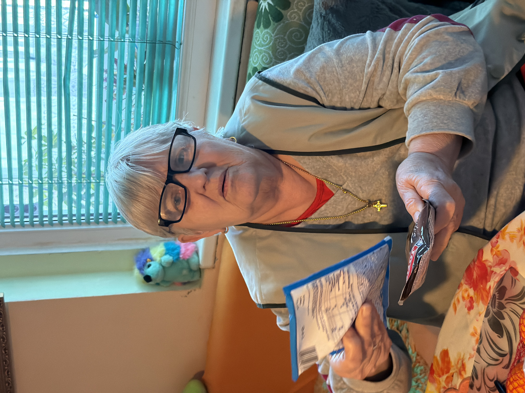 An elderly woman, possibly with short, light grey hair and black-framed glasses, is featured prominently in an indoor setting. She wears a red shirt under a light grey or beige zippered jacket with a dark stripe, and a gold chain with a gold cross pendant around her neck. Her gaze is directed towards the camera with a neutral to slightly smiling expression.
She is holding two small packets in her hands; one is predominantly blue and white with visible text and a barcode, though illegible, and the other is a darker packet, possibly brown or black, with some red and white text (partially visible as "ACTIVE" or "HERO" and "NUTRI...").
The background features a window with light teal or turquoise vertical blinds, suggesting daylight filtering in. A light-colored windowsill below the window holds a small, colorful plush toy that resembles a hedgehog. A green patterned cushion or pillow is partially visible behind the woman's right shoulder. Part of a floral-patterned fabric, possibly a tablecloth or clothing, is visible in the lower right foreground. The overall setting appears to be a casual home environment, possibly in Varna, Bulgaria.