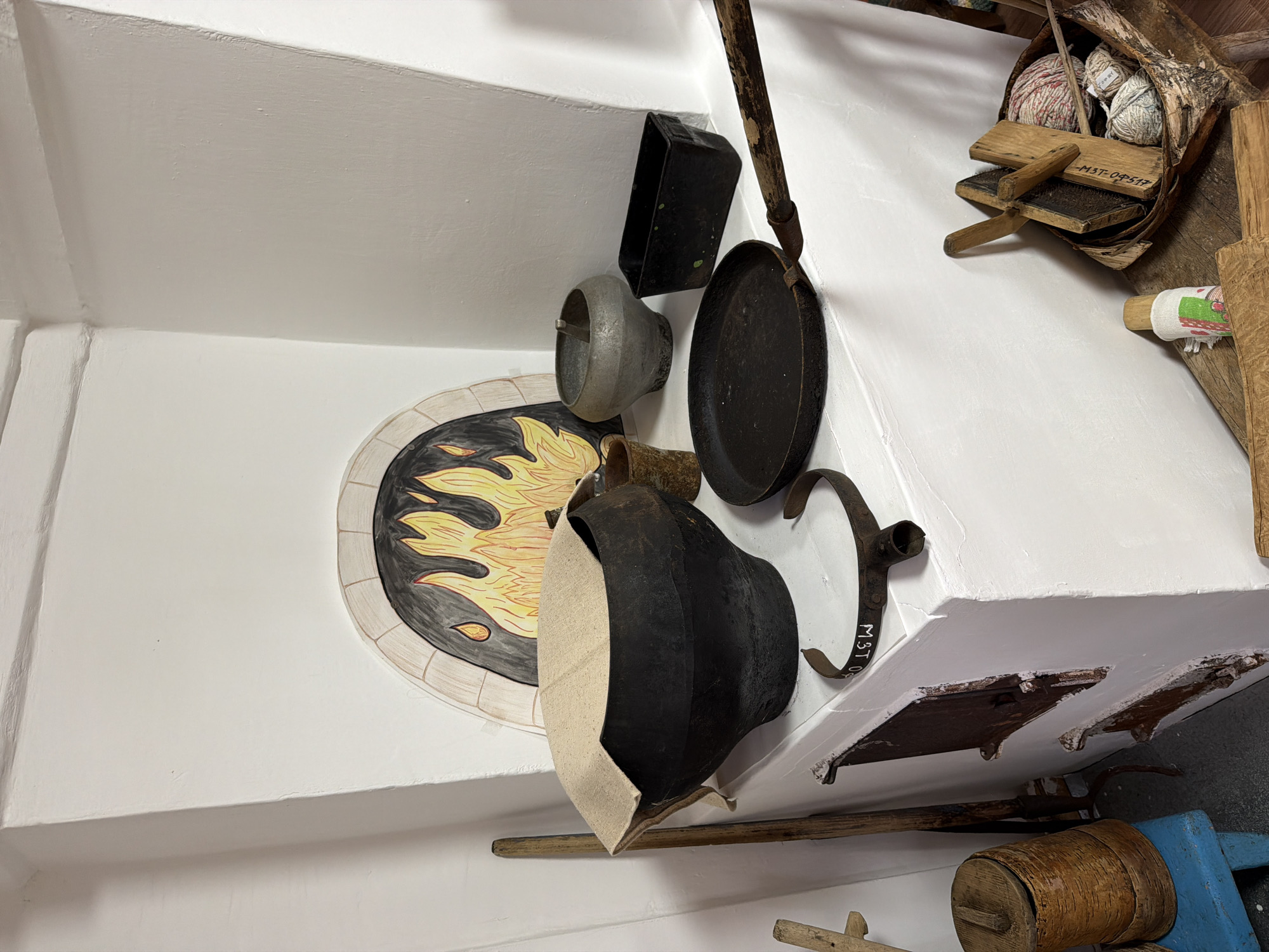 The image depicts a corner of a room with a white stove, likely a traditional Russian stove, against a white wall. The stove has a painted archway resembling a fire with flames. On top of the stove, there are several objects related to cooking and traditional crafts. A large, black, cast-iron pot, partially covered with a light-colored cloth, sits prominently in the foreground. To its right, there is a smaller, grey, metallic pot, and to the right of that, a black rectangular metal container. In front of the pots, a black frying pan with a wooden handle is placed on the stove's surface. A U-shaped metal clamp with white markings is also visible on the stove.
To the left of the stove, a tall wooden stick leans against the wall. Below the stove, there are two rusty metal doors, suggesting an oven or ash compartment. To the far left, a small blue wooden stool holds a cylindrical wooden container with a lid.
In the right foreground, a woven basket rests on a wooden surface. Inside the basket are several balls of yarn, various wooden tools, and what appears to be a bundle of natural fibers. A small doll-like object with a colorful fabric wrap is also present near the basket.
The scene suggests a historical or ethnographic display, possibly in a museum or a preserved traditional home. The location is identified as Ust'-Pyzas, Russia. There are no people visible in the image. The lighting is even, illuminating the objects clearly. The overall impression is one of rustic simplicity and historical preservation.