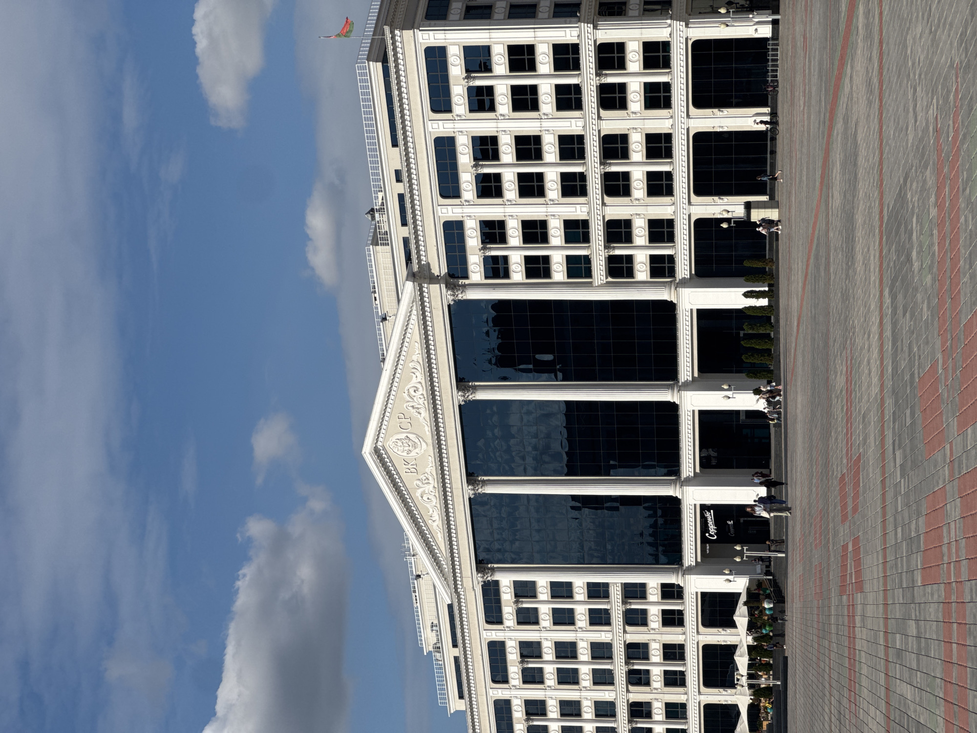 The image shows a large, modern building with a classical facade, located in Minsk, Belarus. The building features a prominent central section with large, dark glass windows framed by white columns, and a triangular pediment at the top. The upper floors have rows of smaller, rectangular windows, also framed by decorative white elements. A flag, red and green, is visible on the roof on the right side of the building. In front of the building is a wide, paved plaza with a pattern of grey and red tiles. Several people are walking across the plaza and near the building entrance. Some are gathered at outdoor tables with umbrellas near the left side of the building, possibly at a cafe. The sky is blue with scattered white clouds. The overall scene depicts a public space with a grand architectural structure.