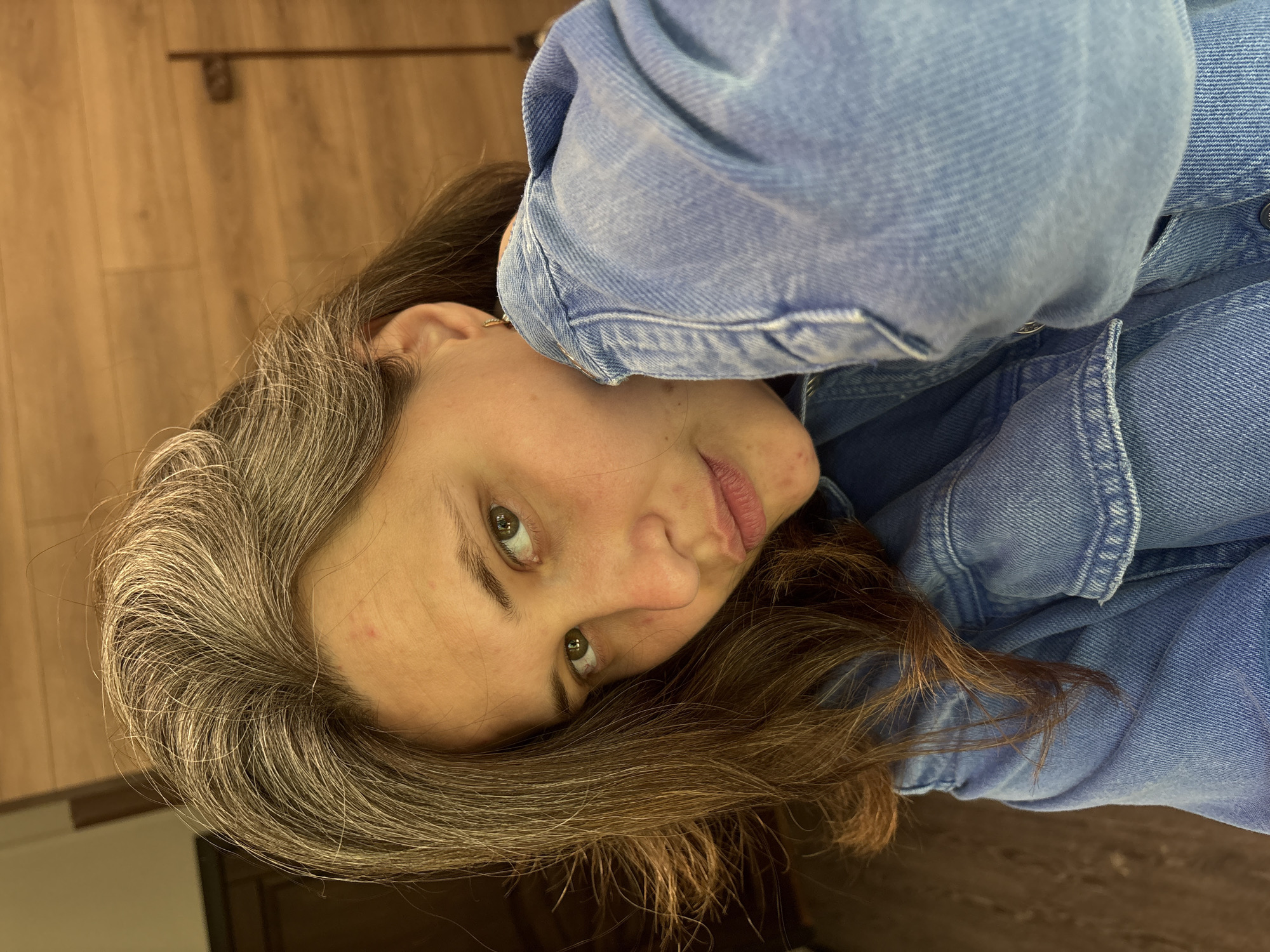A medium shot captures a young woman in a denim jacket from the chest up, her head tilted and angled towards the camera. Her long, brown hair, streaked with gray, is styled loosely, with strands falling across her face. She has green eyes and a light complexion, with a few small blemishes visible on her forehead. Her lips are slightly parted, and her right hand is brought up to her cheek, her fingers gently touching her jawline. She is wearing a small gold earring in her visible right ear. The background appears to be an indoor setting with wooden paneling on the wall behind her. There is no indication of Chaikovsky, Russia, or any specific urban location.