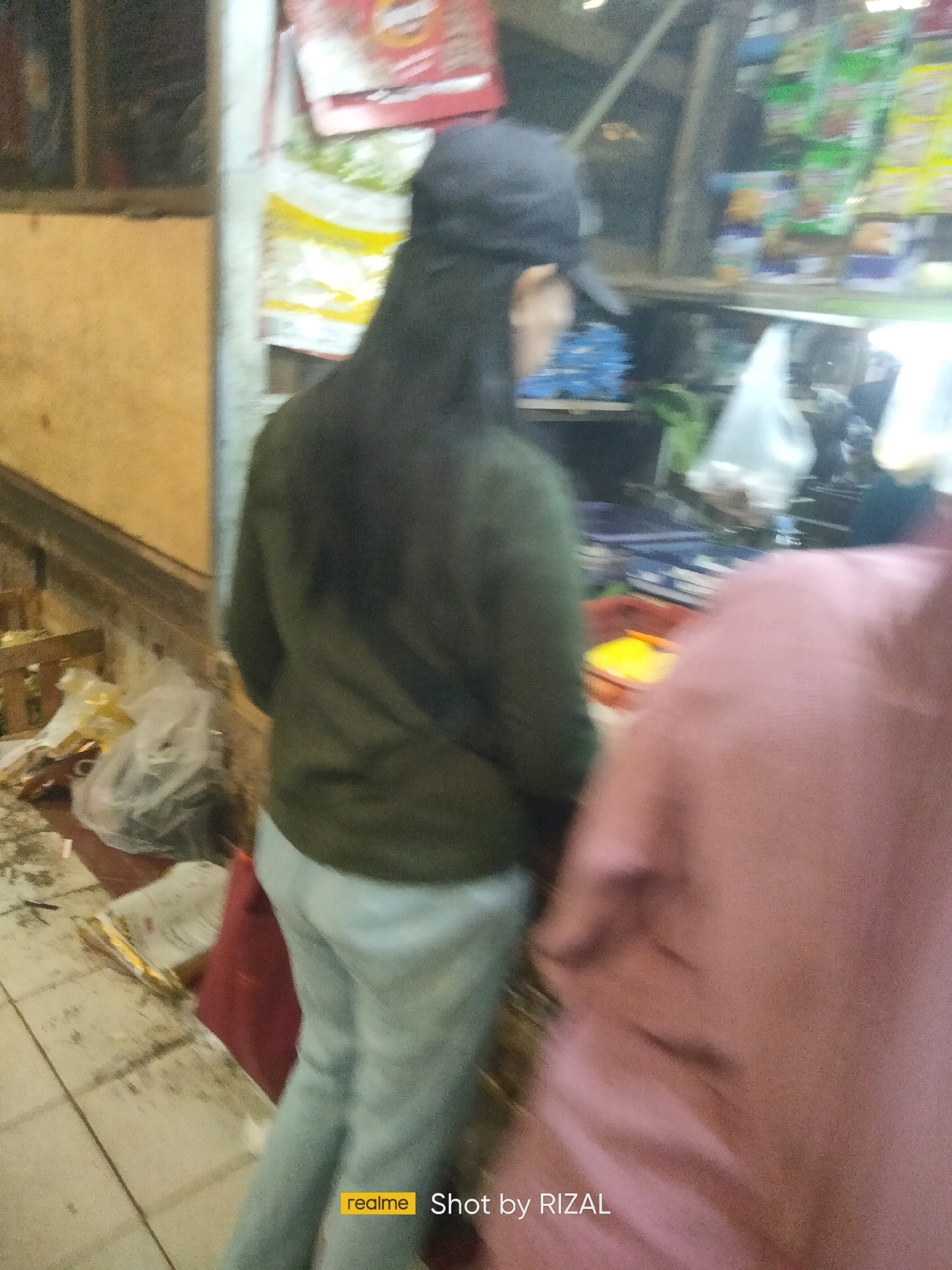 The image depicts a street-level view of what appears to be a market stall or small shop in Bojong Nangka, Indonesia. In the foreground, a person wearing a dark-colored baseball cap and a green long-sleeved shirt is standing with their back to the camera. They are wearing light-colored pants. To the right of this individual, another person's shoulder and upper back are visible, covered by a pink garment, also facing away from the camera. The background features shelves stocked with various items, including packaged goods in green and yellow bags, and what appear to be blue crates or boxes. There are also plastic bags containing items, possibly food or merchandise, and a red bag on the floor. The environment suggests a bustling, possibly outdoor, commercial area with visible shelving units and a tiled floor. The overall impression is a candid snapshot of daily life in a marketplace.
