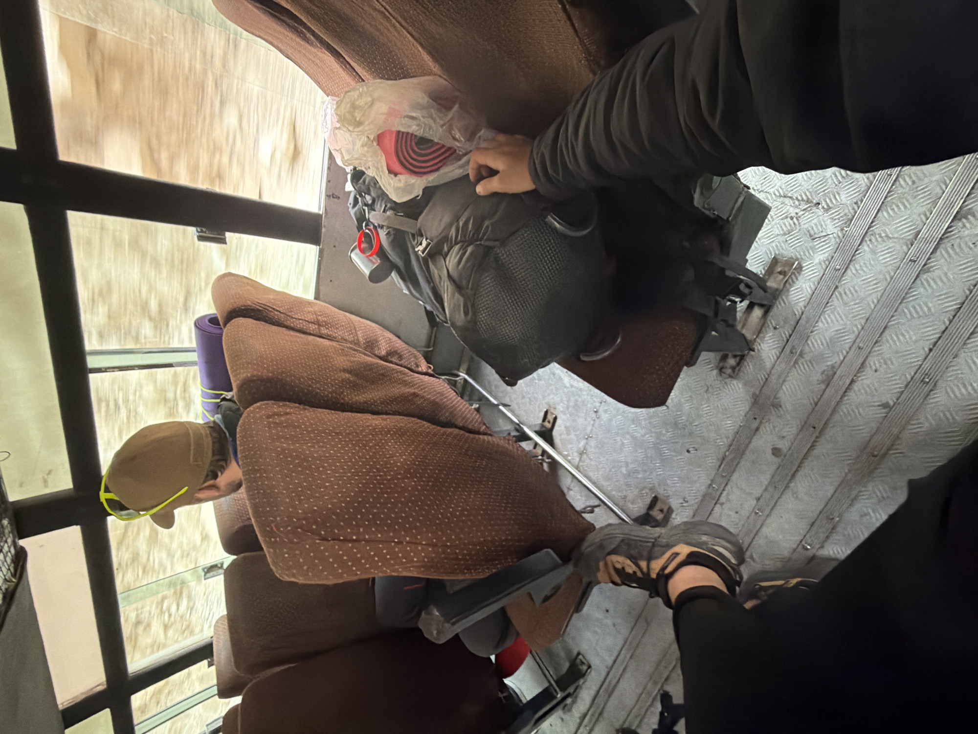 The image depicts the interior of a vehicle, likely a bus, with seats and luggage. In the foreground, a person's legs and foot wearing hiking boots and black pants are visible, suggesting they are a passenger. To the right, a backpack is placed on a seat, with a rolled-up sleeping mat in a plastic bag on top. Another person, wearing a brown baseball cap with neon yellow detailing, is seated in front and looking out the window. The background shows a blurred view through the windows, indicating movement. The flooring of the vehicle is a ridged metal. The scene appears to be in the city of Gayk, India, given the provided location context.