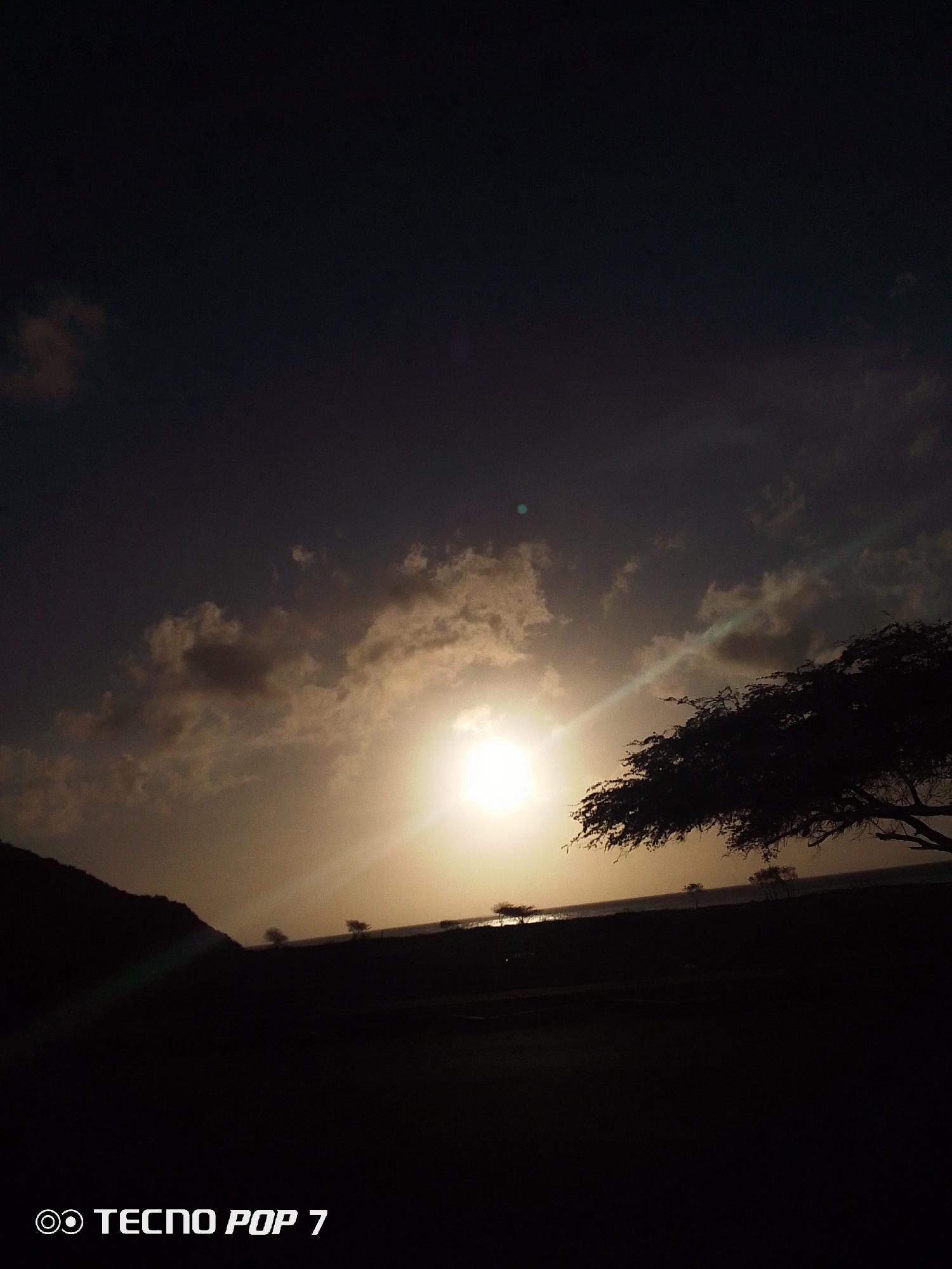 A low-angle shot shows a sunset in Pedro González, Venezuela. The sun is positioned in the center of the frame, obscured by clouds. The horizon is lined with silhouetted trees and a small hill on the left side of the frame. In the foreground, the ground is dark and indistinct. The "TECNO POP 7" watermark is visible in the lower left corner.