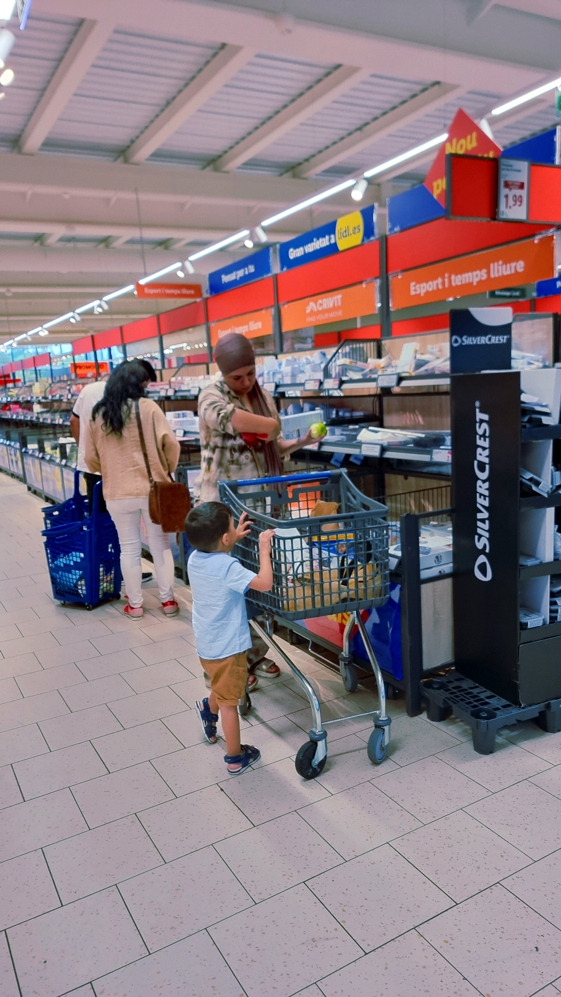 In a brightly lit supermarket aisle in Olot, Spain, a young boy, about four years old, with dark hair, is seen from the waist down, wearing a light blue t-shirt and tan shorts, pushing a grey shopping cart. His hands are on the handle of the cart, and he is wearing dark blue sandals. The cart contains various grocery items. To the right of the boy and slightly behind him, a woman wearing a patterned headscarf and a patterned shirt is seen from the chest up, holding a green apple. She appears to be interacting with the cart. Further back and to the left, a woman with dark hair, wearing a tan cardigan over a white top and white pants, is walking away from the camera, pushing a blue shopping basket. The supermarket aisle has beige tiled flooring. Above the aisle, advertising banners are visible, including one that says "Gran varietat a lidl.es" and another that says "Esport i temps lliure". A display stand labeled "SILVERCREST" is also visible.