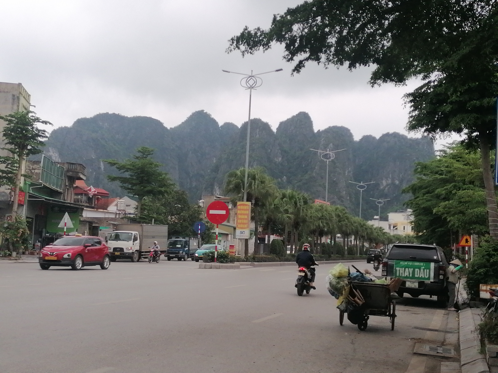 This image captures a street scene in Ha Tu Ward, Vietnam, under an overcast sky, with dramatic karstic limestone mountains forming a prominent backdrop. The light is soft and diffused, suggesting a cloudy day, possibly morning or late afternoon.
In the foreground, a paved road stretches across the frame, indicating an active street or intersection. Several vehicles are present: a red crossover SUV (possibly a Hyundai Kona) is visible on the left, appearing to turn; further down, a white truck and a blue truck are parked or moving slowly. Two motorcycles are also seen on the road, one in the mid-ground and another further back. On the right side of the road, a dark green or black pickup truck is parked by the sidewalk. A person, wearing a conical hat and gloves, is sweeping the sidewalk next to this truck. Nearby, a two-wheeled cart laden with various items, possibly refuse or recyclables, is being pushed by an unseen person or is stationary near the road.
The street is lined with buildings, primarily low-rise structures with visible rooftops and some storefronts. Palm trees are planted along the median or side of the road, and other leafy trees provide shade. Distinctive streetlights with circular, ornate designs are visible in the mid-ground, extending down the street.
Notable street signs include a circular red "do not enter" sign on the median and a yellow sign with "5G Mobifone" partially visible. The pickup truck prominently displays Vietnamese text: "DỊCH VỤ (SERVIE)" and "THAY DẦU", which translates to "Service" and "Oil Change". The overall scene conveys the bustling yet calm atmosphere of everyday life in a Vietnamese town, set against a striking natural landscape.