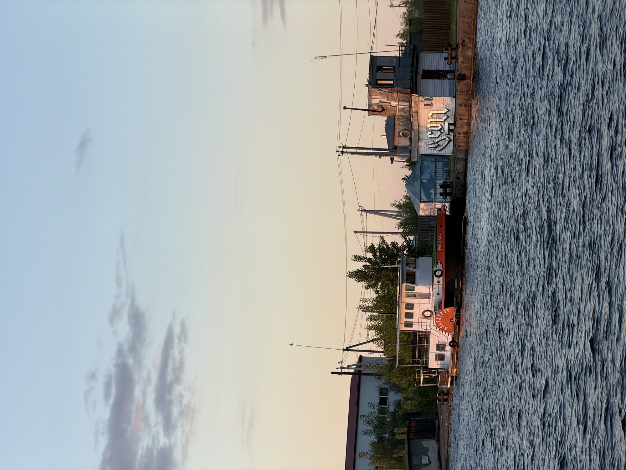A long shot captures a canal in the city of Chaikovsky, Russia, at dusk. The sky displays a gradient of soft blues and pinks, with a few wispy clouds. The water in the foreground is dark and rippled, reflecting the twilight.
Along the canal bank, several structures are visible. On the left, a long, white building with a red roof and windows stands behind a line of trees. Next to it, a white boat with a red stripe and the name "АЛЬБАТРОС" (Albatross) in red letters is docked. The boat has a cabin and a railing along its deck. A life preserver is visible on its side. Further down the bank are several utility structures. One is a tall, weathered building with a control tower-like structure on top, adorned with graffiti. Another is a smaller, blue tent-like structure.
Power lines crisscross the scene, supported by utility poles. The overall atmosphere is calm and serene, typical of a quiet evening in a port town. There are no people or animals visible in the frame. The scene suggests a functional industrial area combined with the natural beauty of the canal and the sky.