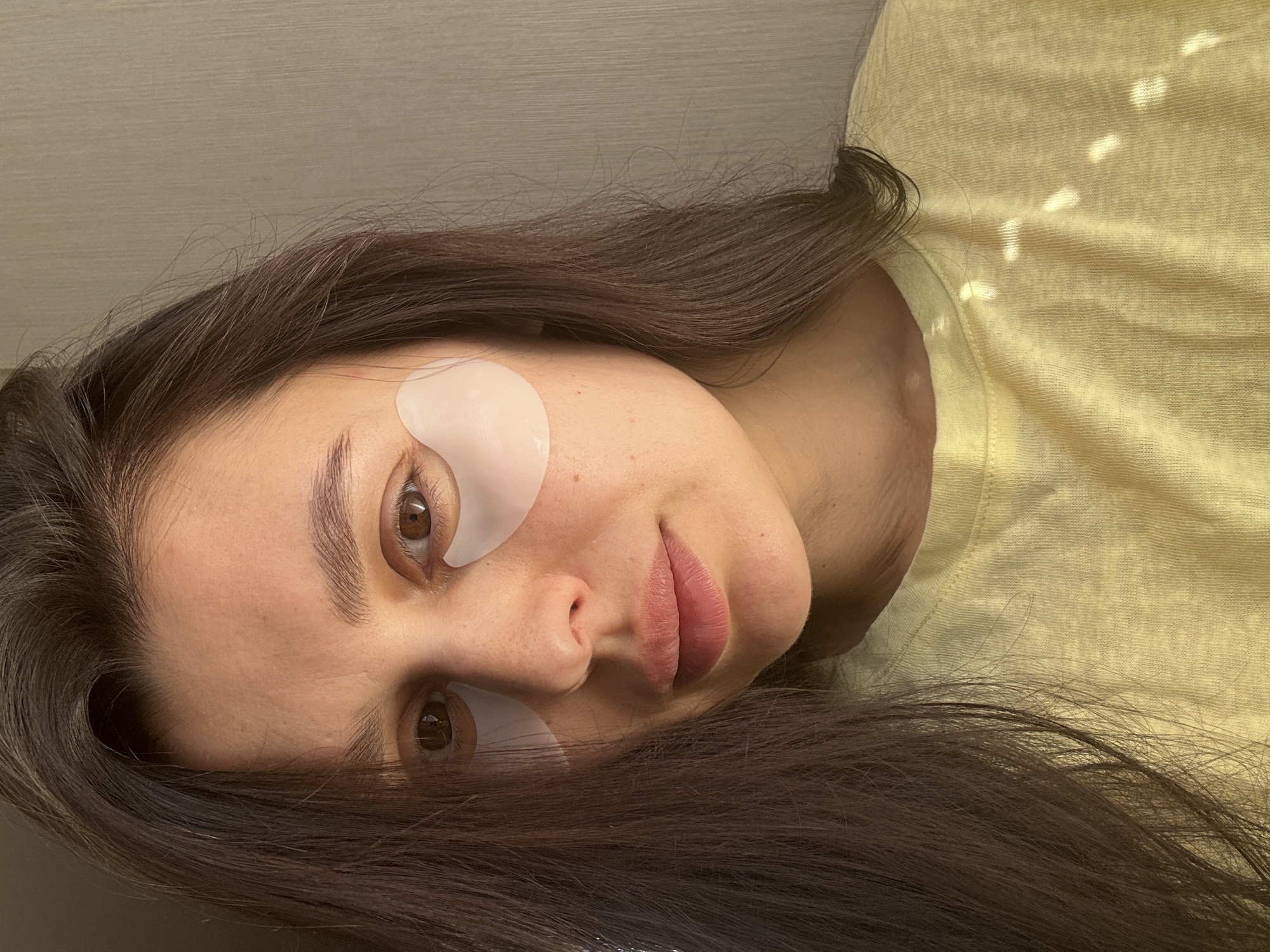 A young woman with dark brown hair is looking at the camera. She has fair skin and brown eyes. Under each eye is a white, crescent-shaped eye patch. She is wearing a light yellow T-shirt. The background is a light tan color. It is not possible to determine the location of the photo from the given image.